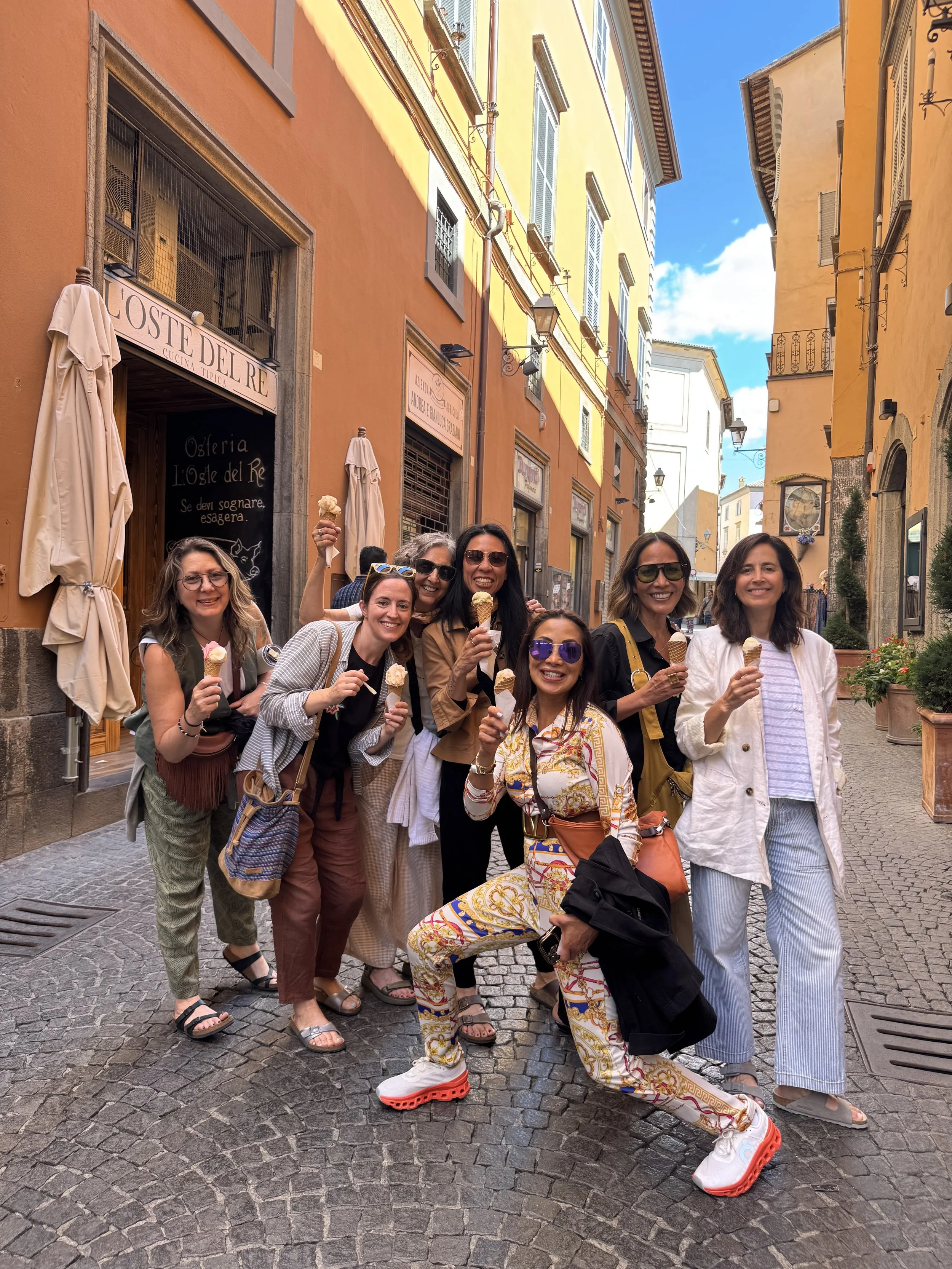 A group of seven women standing on a cobblestone street in front of colorful European-style buildings, smiling and holding ice cream cones, enjoying a sunny day.