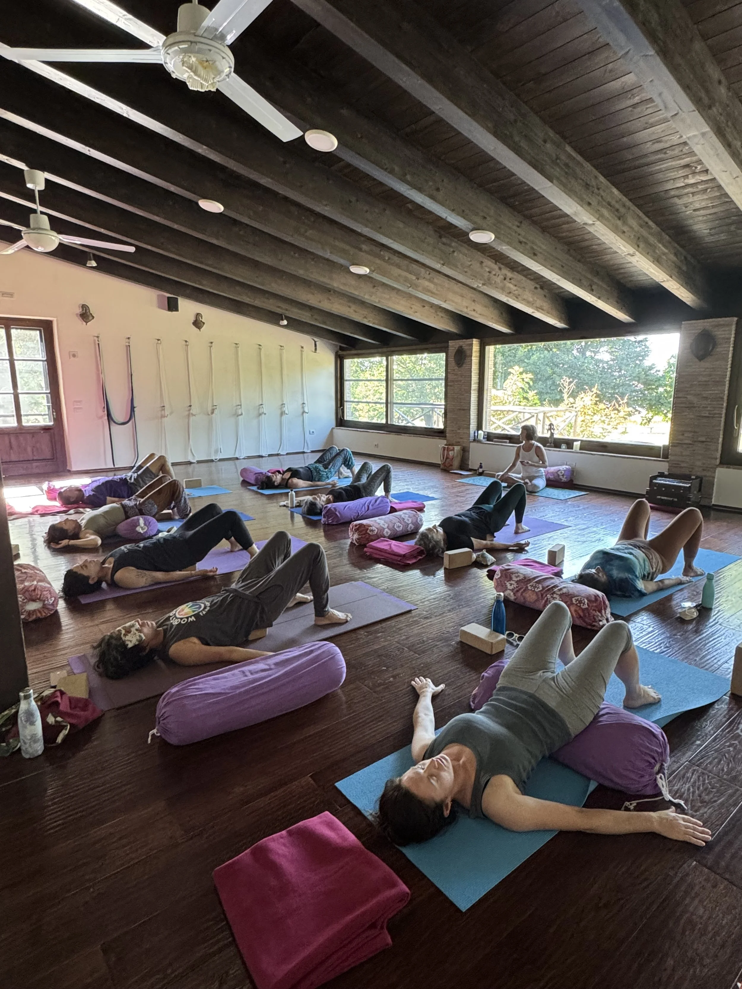 People participating in a yoga class or meditation session in a rustic wooden-ceiling studio with large windows, practicing relaxation poses on mats with cushions and water bottles, led by an instructor.