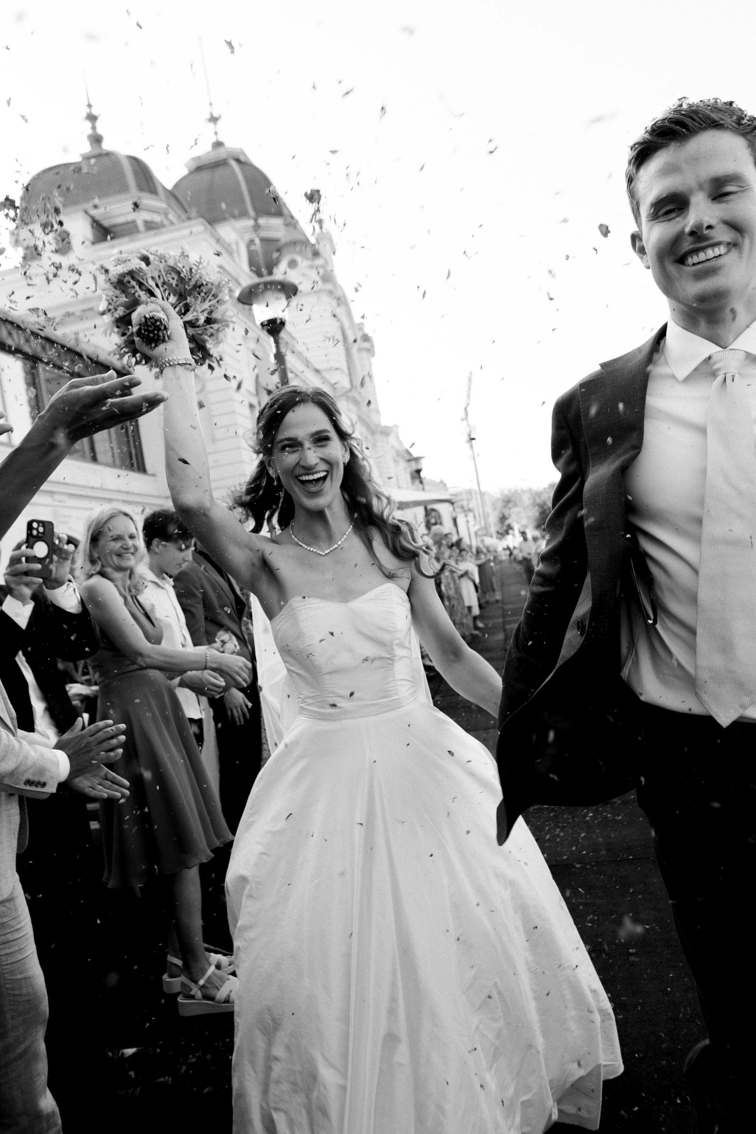 A happy couple is dressed in wedding attire and celebrating outdoors with guests around them. The bride in a white wedding dress is smiling and holding out her arm, while the groom in a suit is smiling as well. There is confetti in the air and a buil