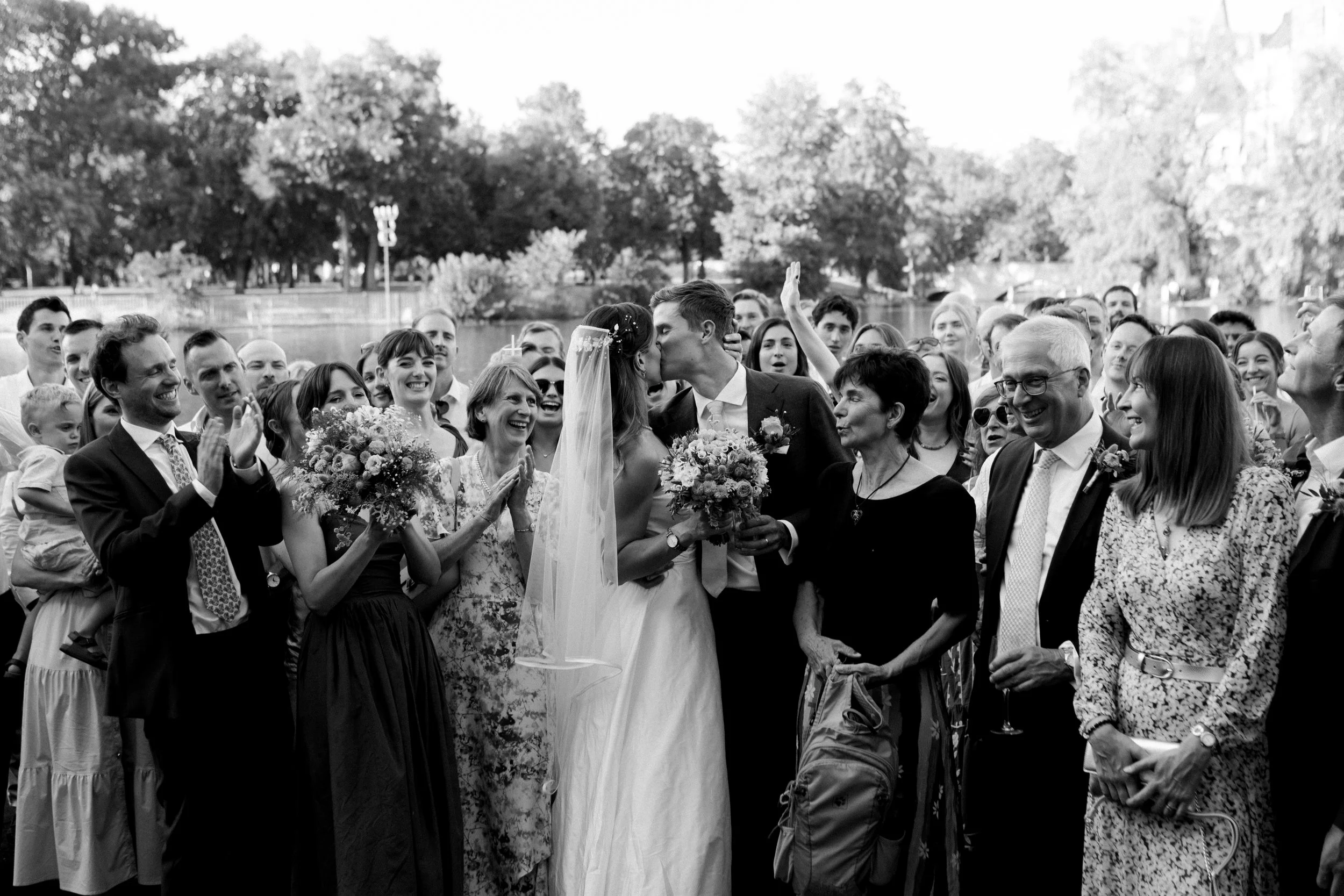 A wedding celebration outdoors by a lake with a large group of people gathered around the bride and groom. The bride and groom kiss while holding bouquets, and the guests smile, clap, and cheer.