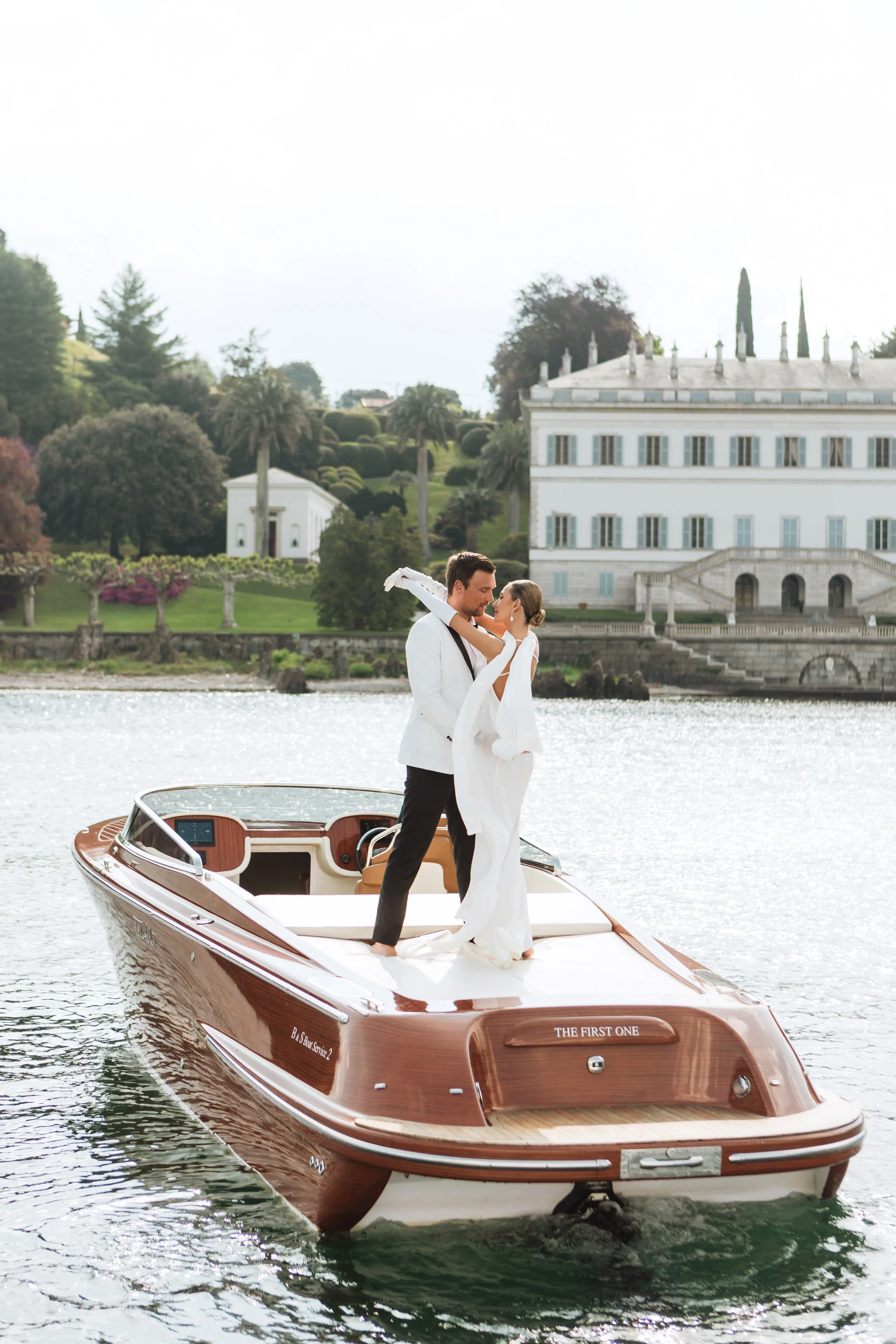 Wedding portrait shoot, where a couple dressed in wedding attire dances on a yacht in a body of water with a grand white mansion and landscaped greenery in the background at Lake Como.