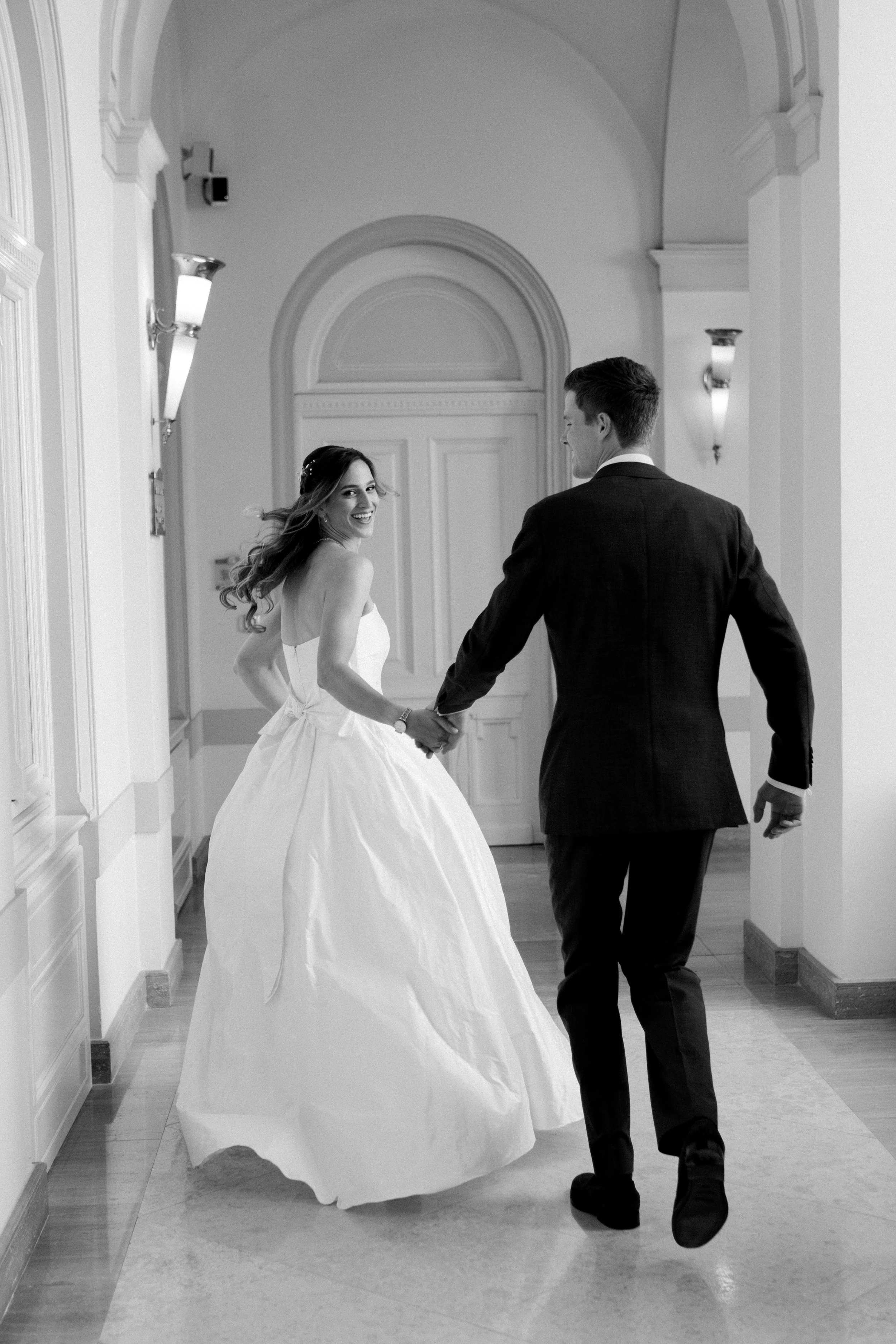 A happy bride and groom holding hands and walking through a hallway with ornate molding and wall sconces, in black and white.