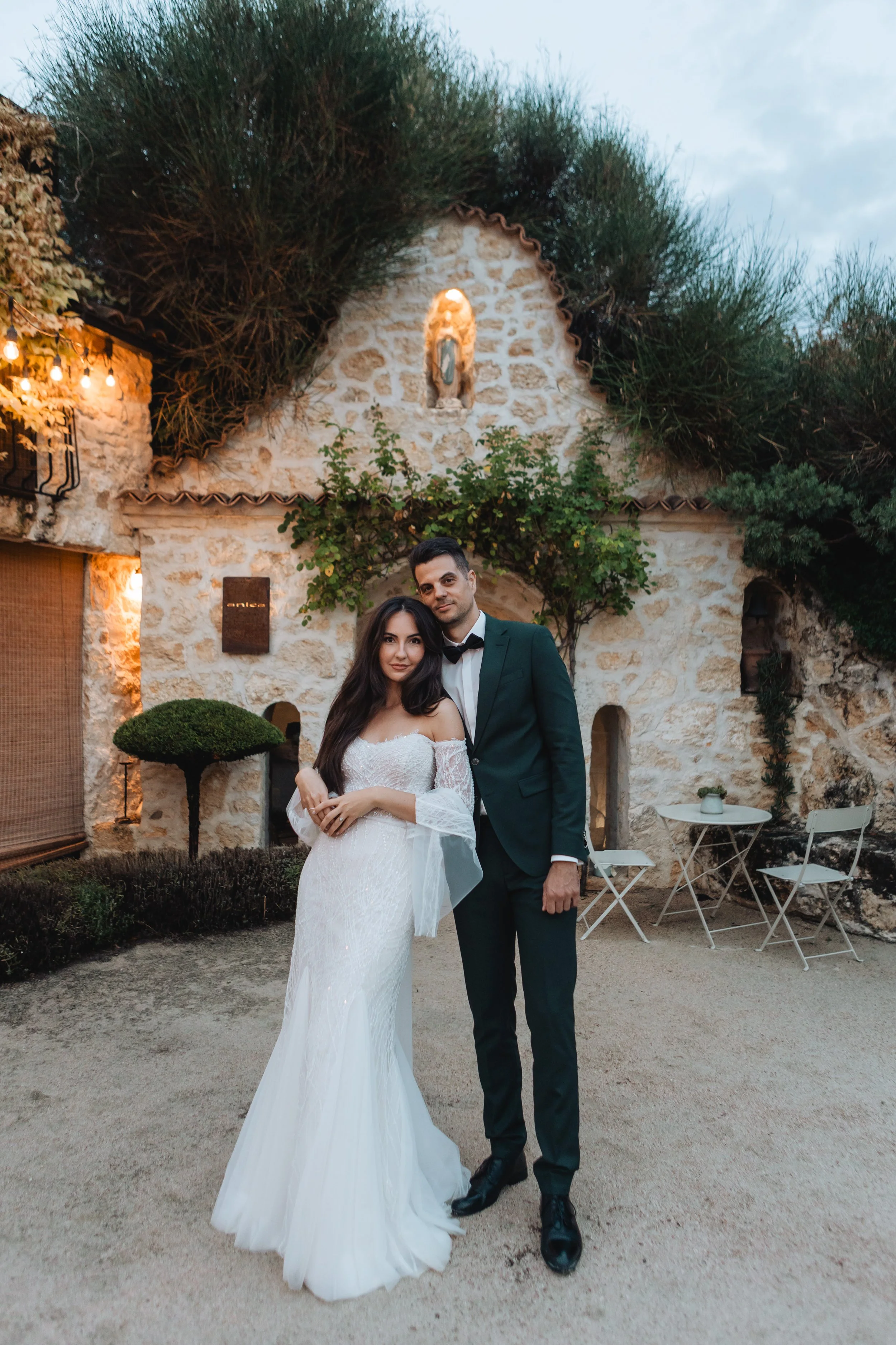 A bride and groom stand together outdoors in front of a rustic stone building with a small arched window and greenery. The bride wears a white lace wedding dress, and the groom wears a dark green suit with a bow tie.