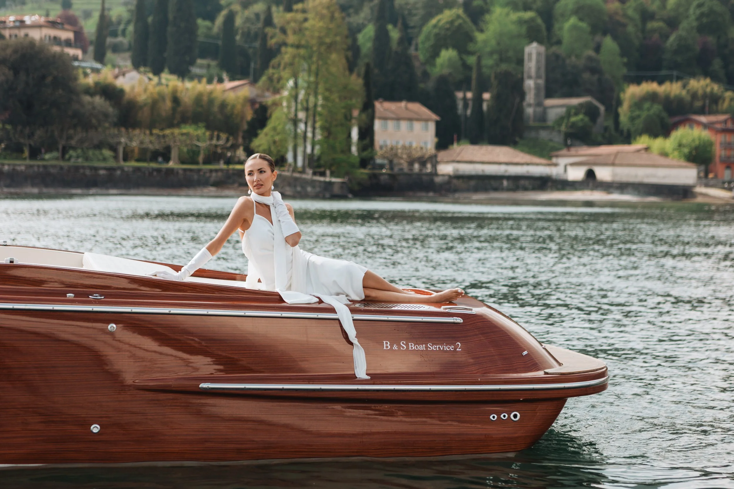 A woman in a white dress and gloves is sitting on the edge of a wooden boat on a lake, with green trees and houses in the background.