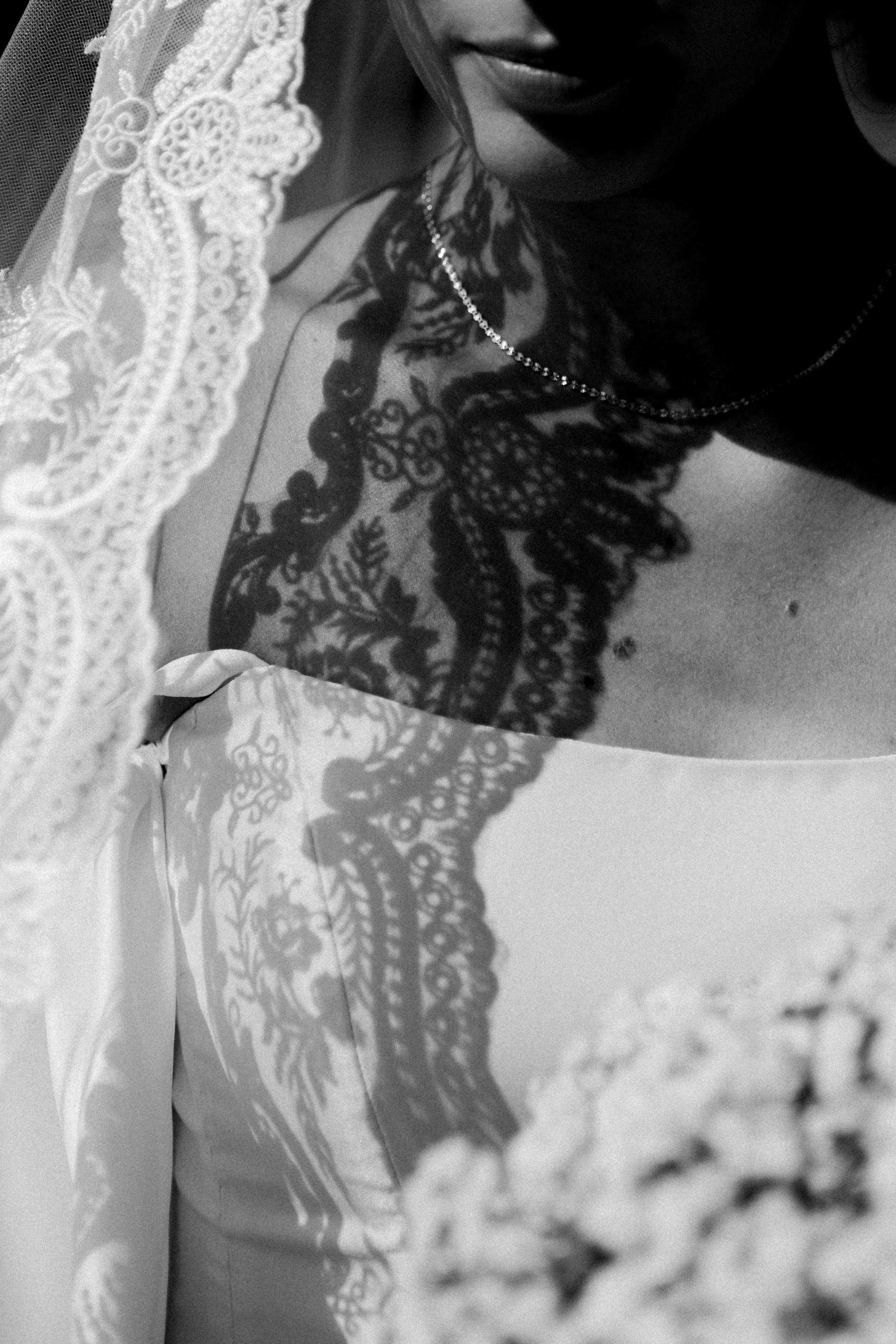 Close-up of a woman wearing a lace veil and a necklace, with intricate lace fabric in the foreground, and her face partially visible in black and white.