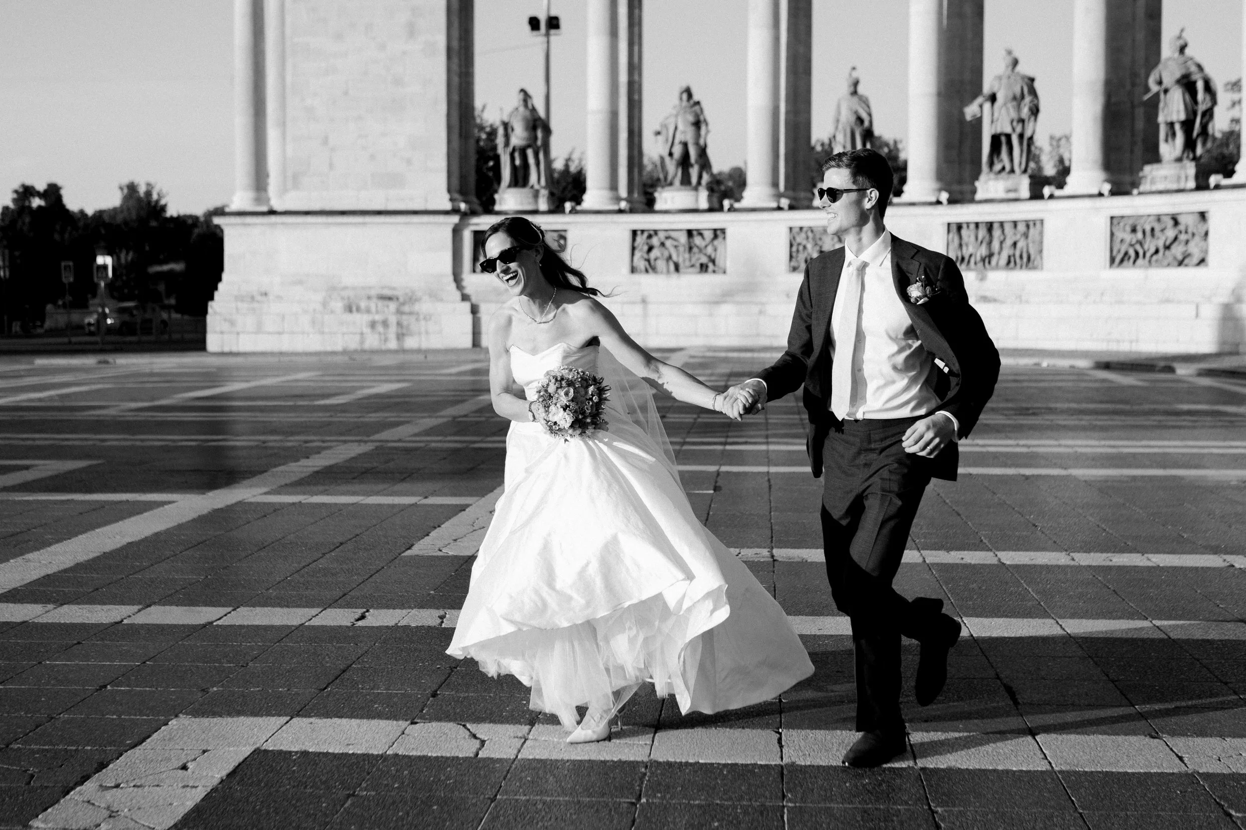 A bride and groom holding hands and walking together, both wearing sunglasses. The bride is in a strapless wedding gown holding a bouquet, and the groom is in a suit with a tie. They are smiling and looking at each other, with a historical monument f