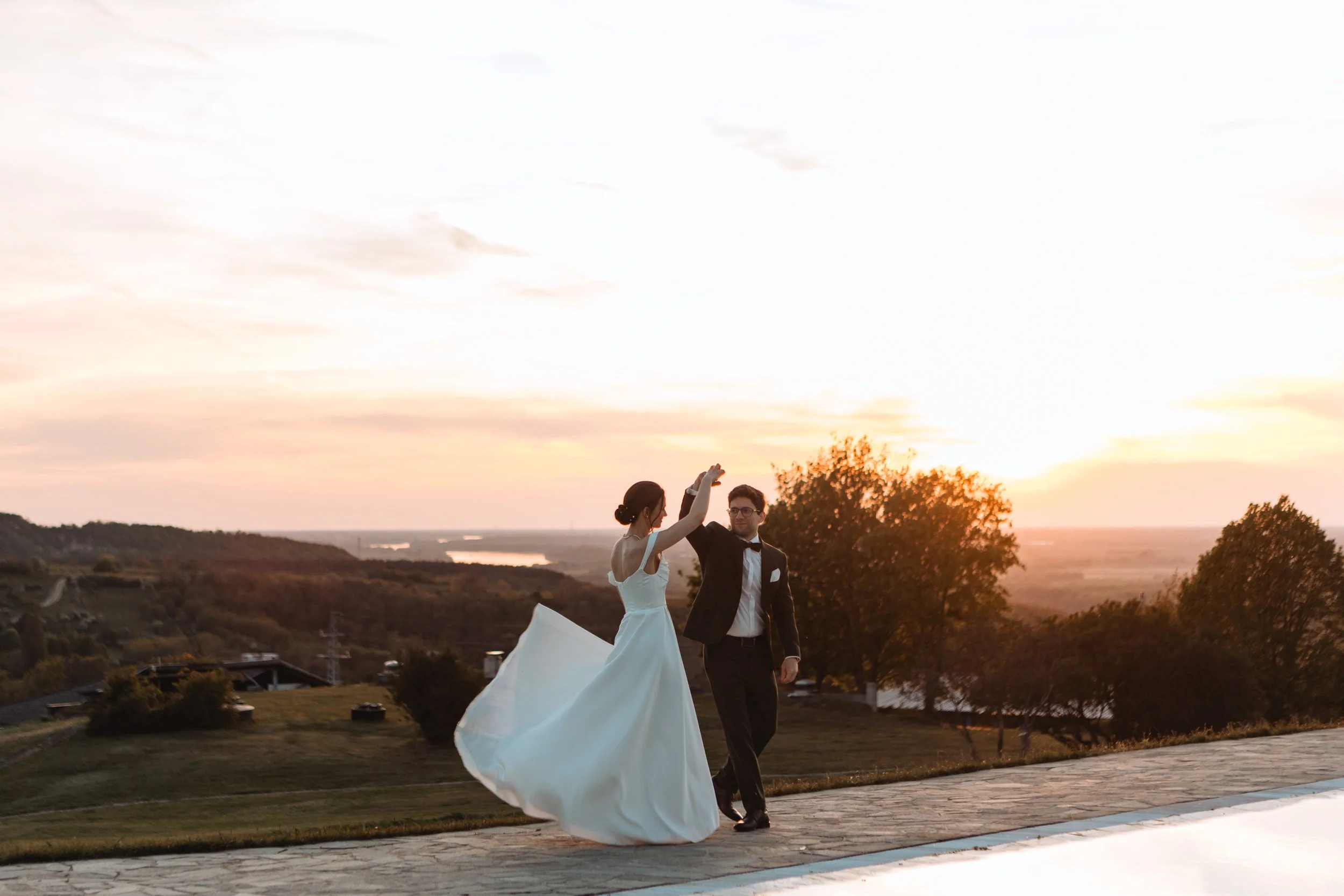 A couple dressed in wedding attire dancing outdoors during sunset, with trees and a landscape in the background.