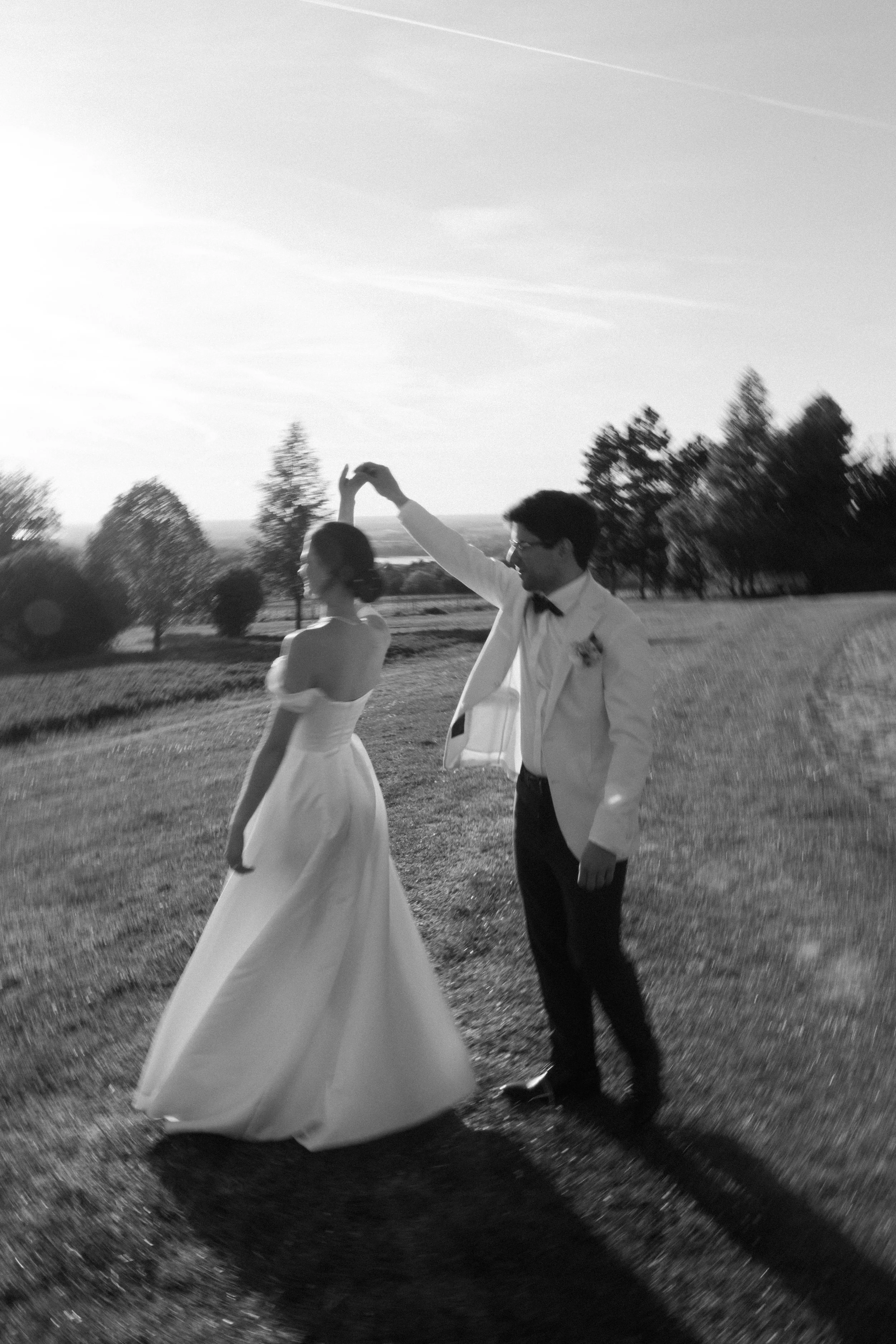 A black and white photo of a newlywed couple dancing outdoors on a grassy field, with trees in the background and a clear sky overhead. The man is wearing a tuxedo with a bow tie, and the woman is in a strapless wedding gown.
