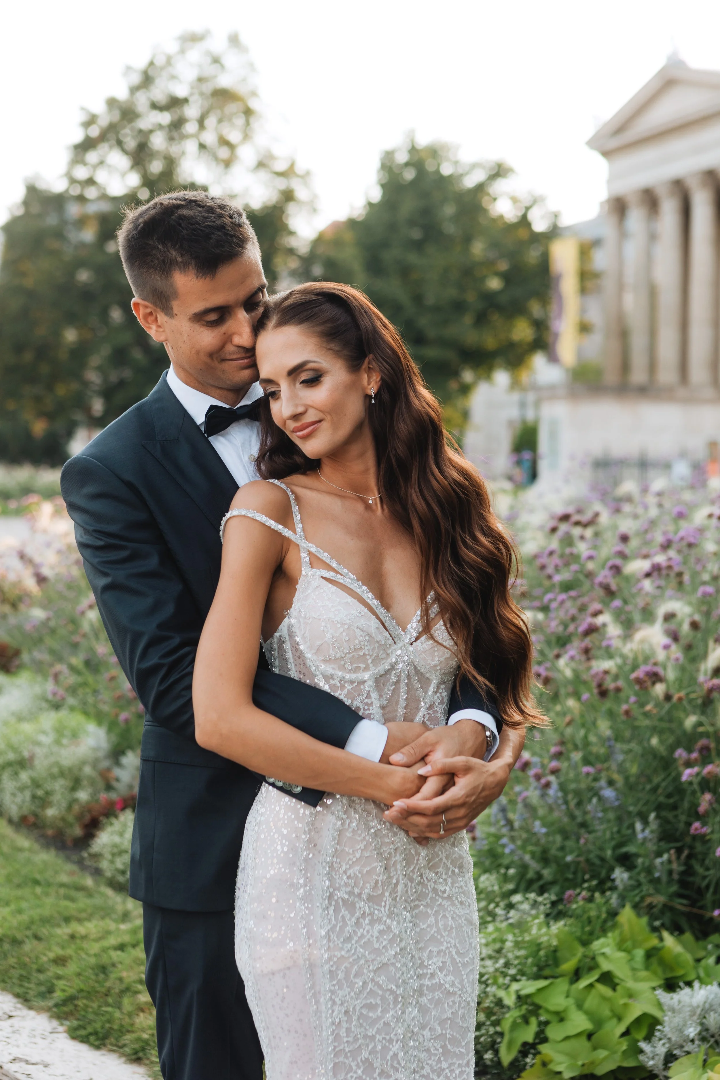 A happy couple in wedding attire standing in a garden filled with flowers, with the groom in a tuxedo and the bride in a white lace wedding gown. The groom is embracing the bride from behind, and they both seem content and loving.