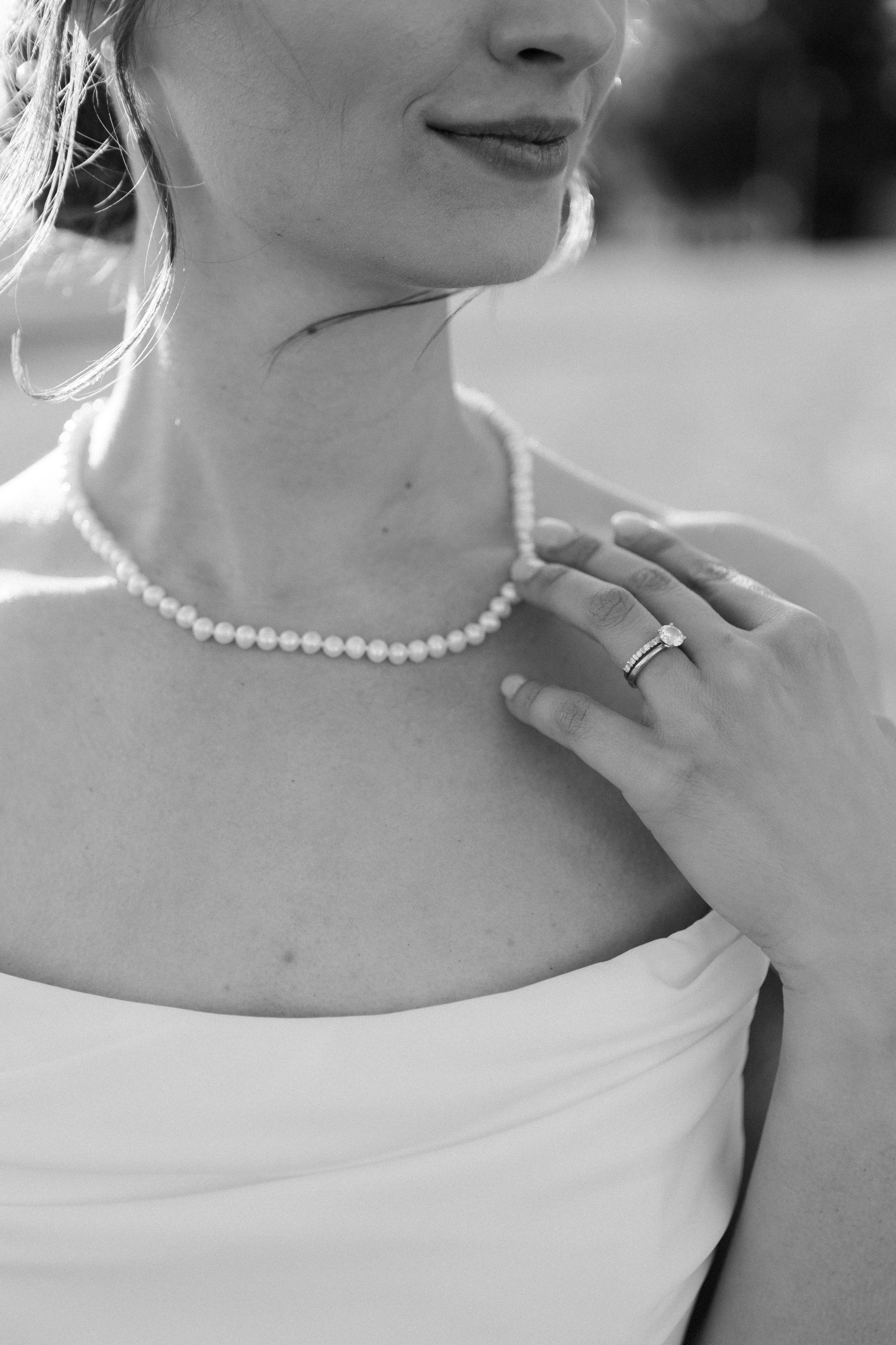 Close-up of a woman wearing a pearl necklace and ring, with her hand on her chest, in a strapless white dress. She has light-colored wavy hair, and her face is partially visible with a slight smile. The background is blurred.