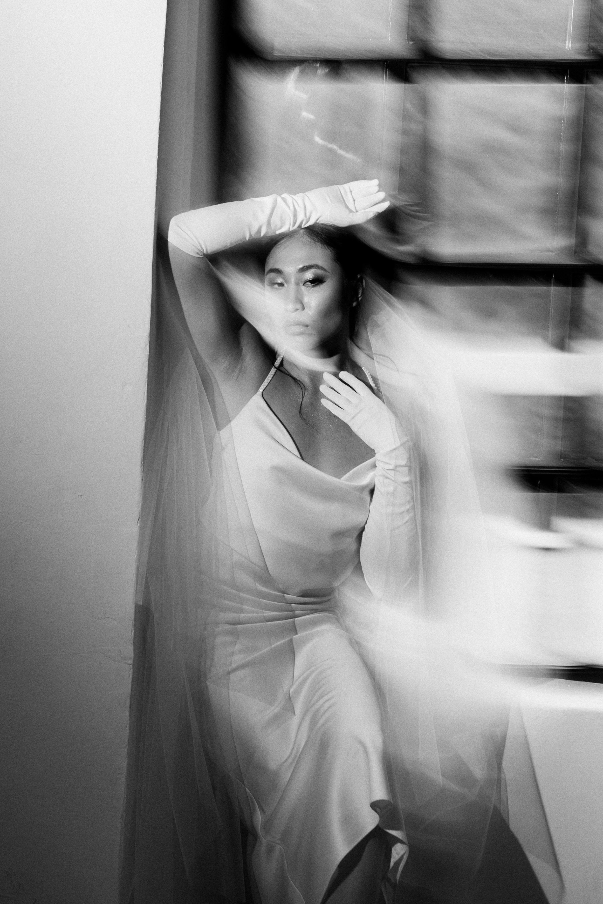 Black and white photo of a woman in a satin dress with long gloves, posing with her arm raised and resting against wall, with a blurred background.