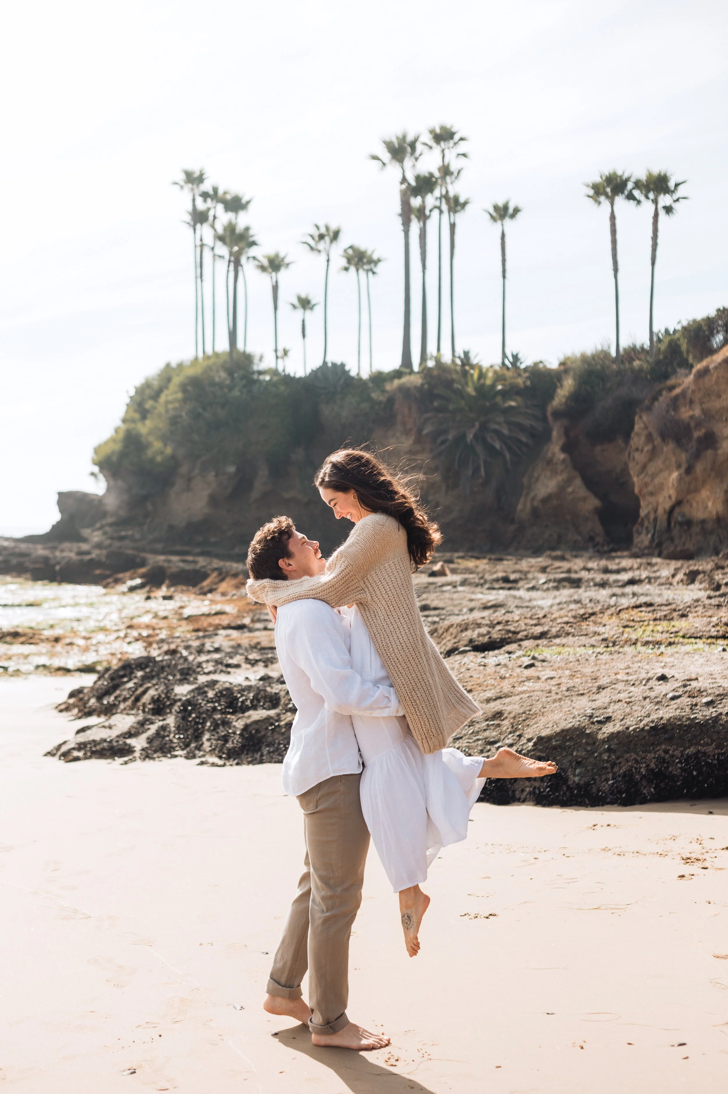 A couple on a beach with a rocky cliff and palm trees in the background in California. The man lifts the woman, and they are smiling at each other. The woman has one leg bent with her foot off the ground, and the man is barefoot.
