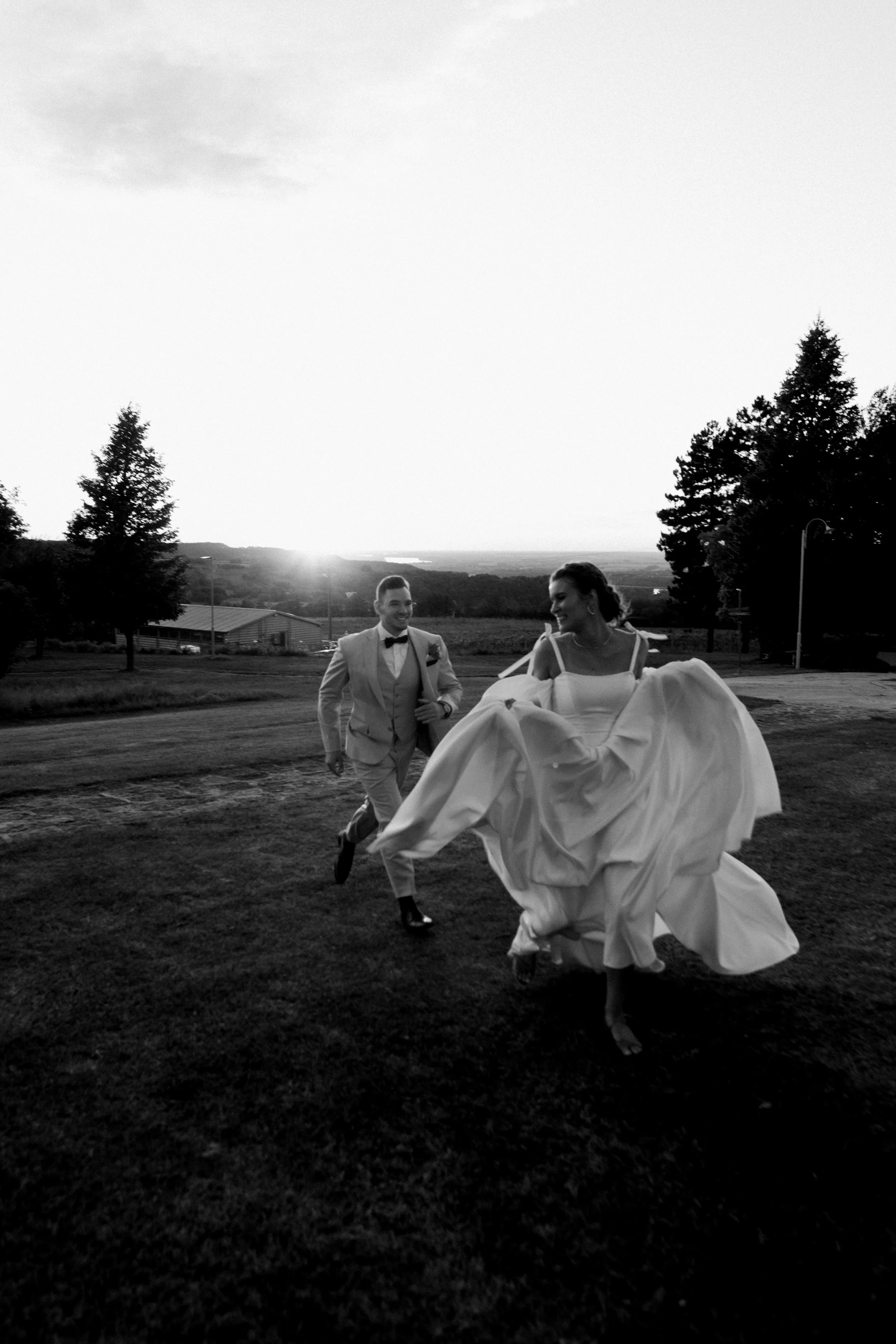 Black and white photo of a couple, with the man in a tuxedo and the woman in a wedding dress, running outdoors during sunset, smiling and enjoying each other's company.
