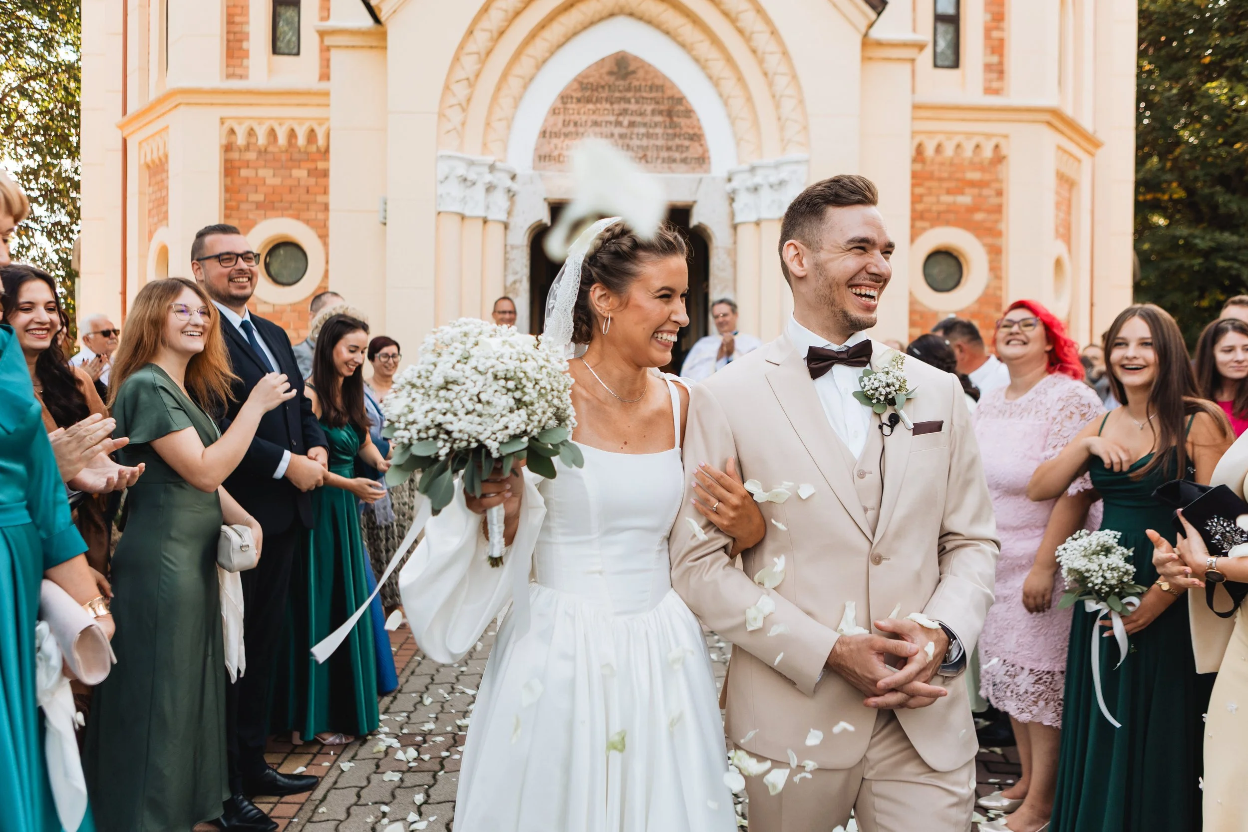 A bride and groom walk outside a church surrounded by their wedding guests. The bride is holding a bouquet of white flowers and smiling. The groom is wearing a light-colored suit with a bow tie. The guests are smiling and celebrating, some clapping, 