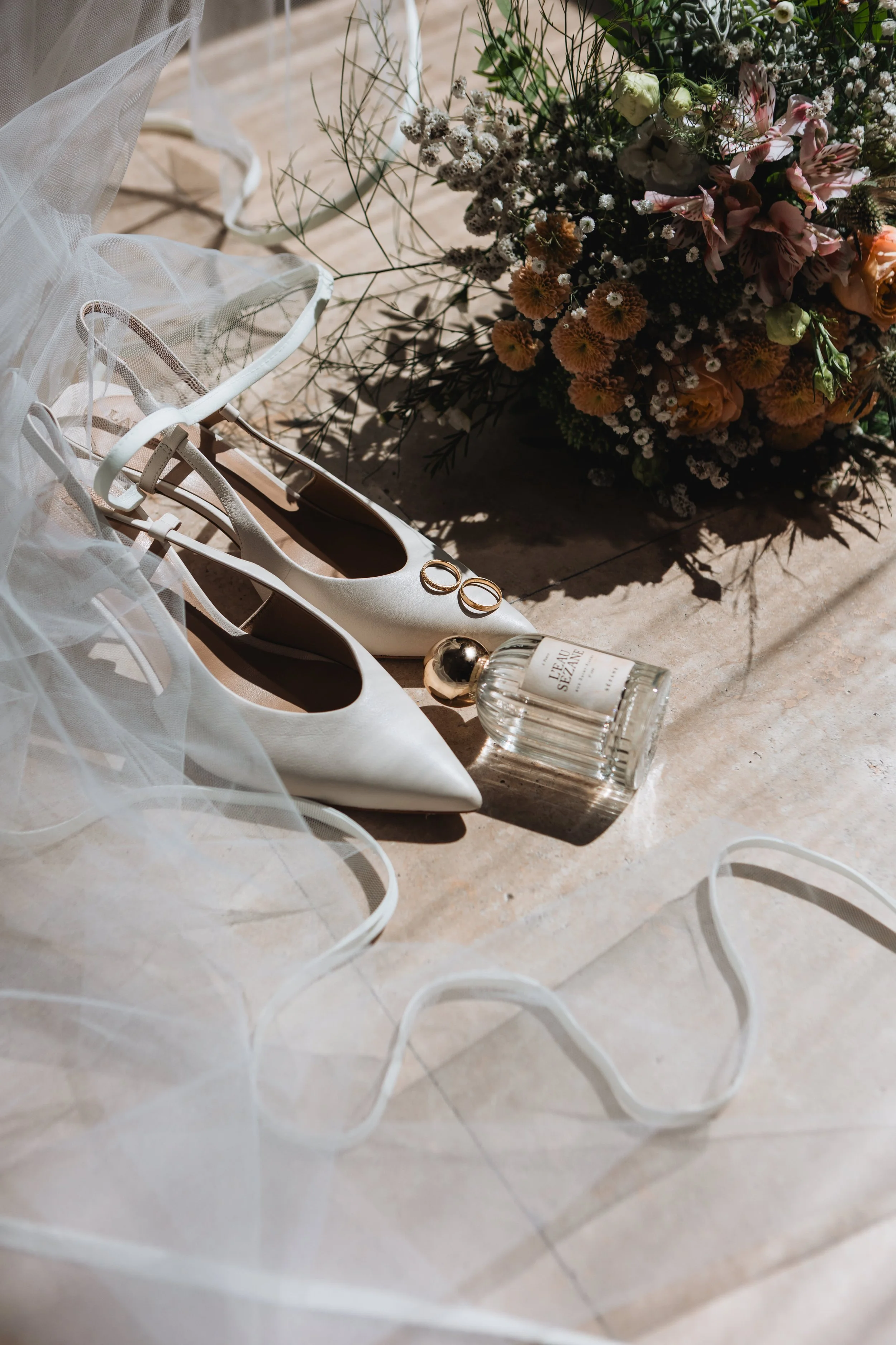 Wedding shoes, a bouquet of flowers, a perfume bottle, and gold rings on a light-colored surface, with sunlight and shadows.