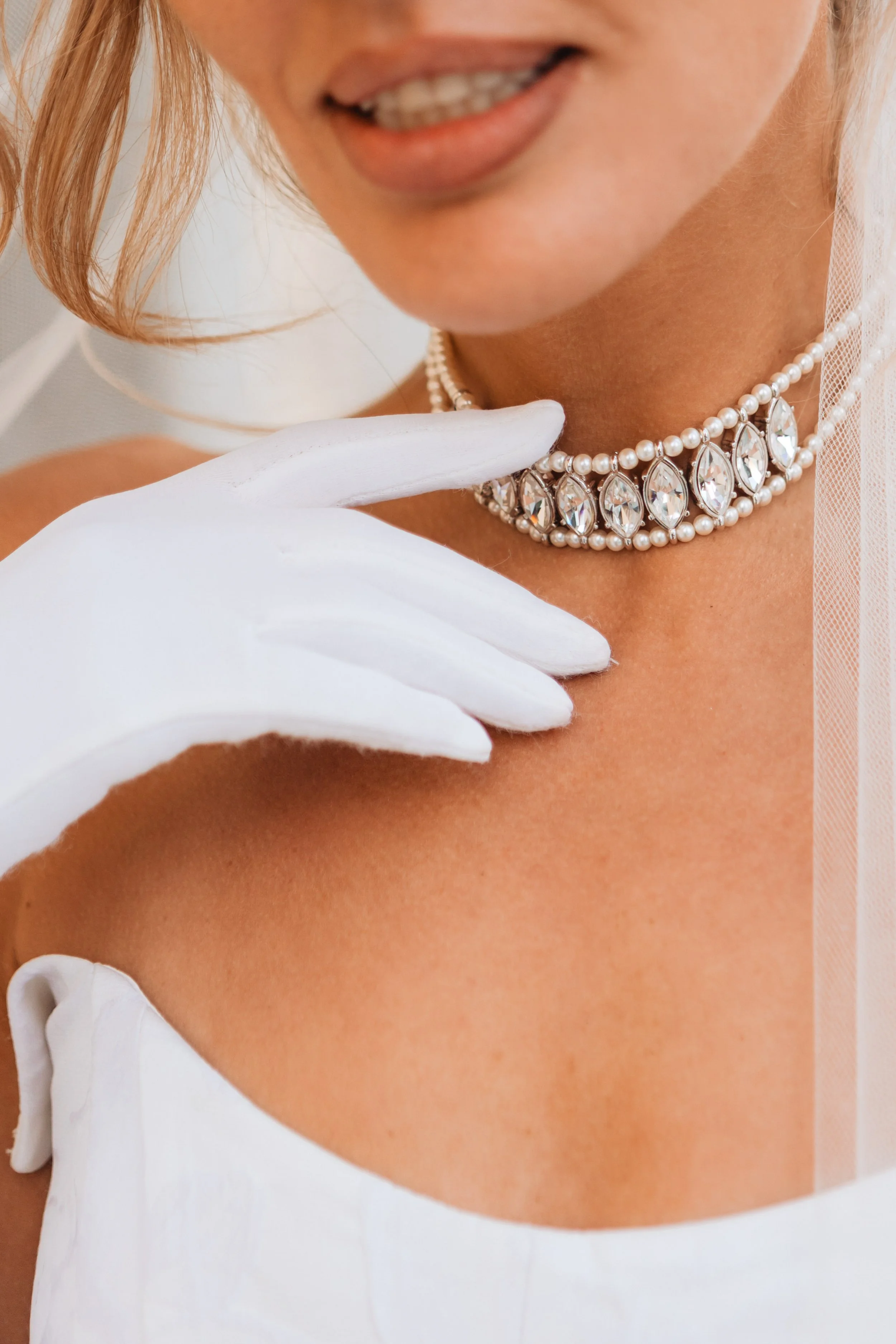 Close-up of a woman wearing a pearl and diamond necklace, white gloves, and a white dress, with her lips slightly parted.