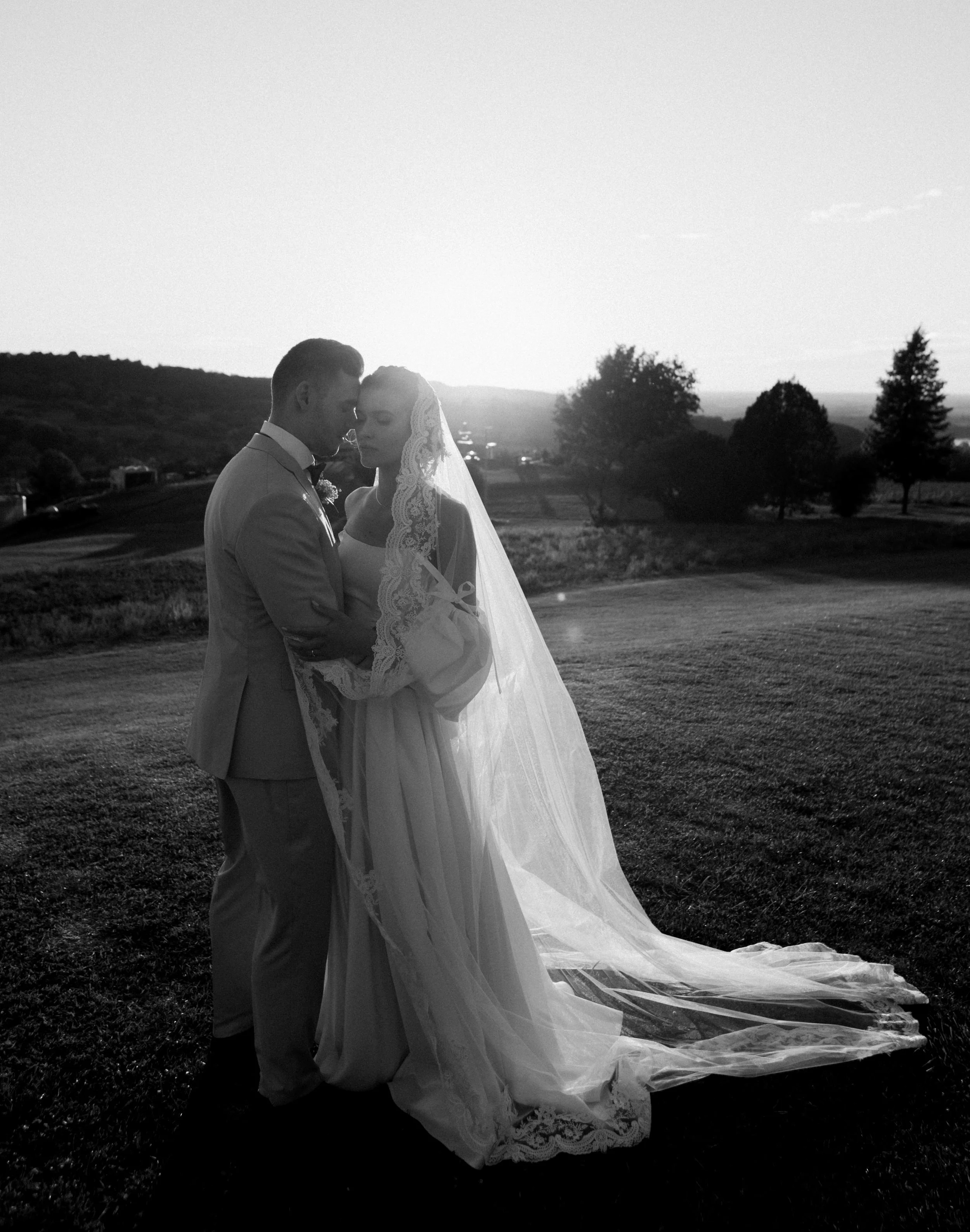 Black and white photo of a bride and groom embracing outdoors during sunset, with the bride wearing a lace veil and gown and the groom in a suit, standing on a grassy field with trees and hills in the background.