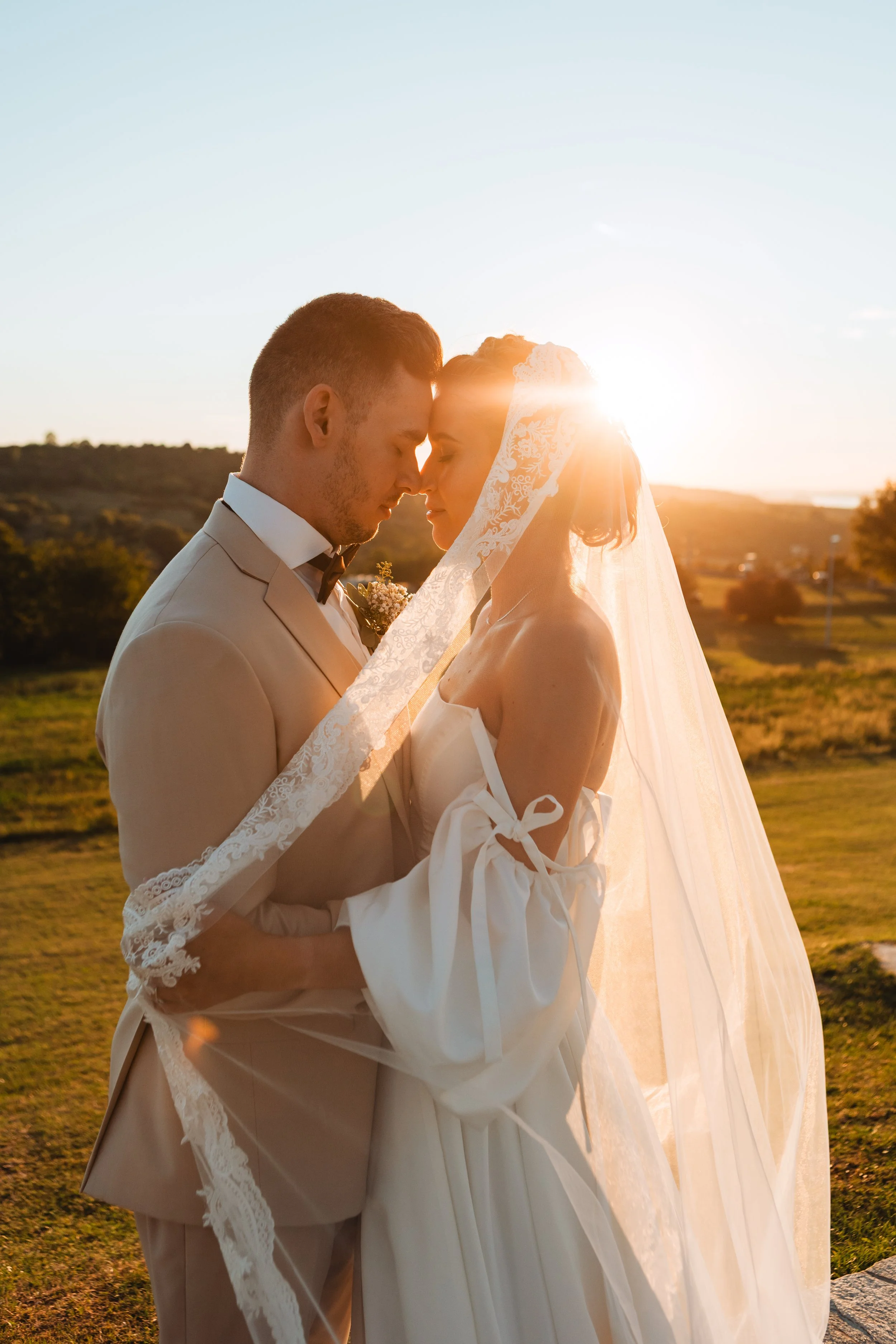 A bride and groom standing close together with foreheads touching during sunset, in a scenic outdoor setting.