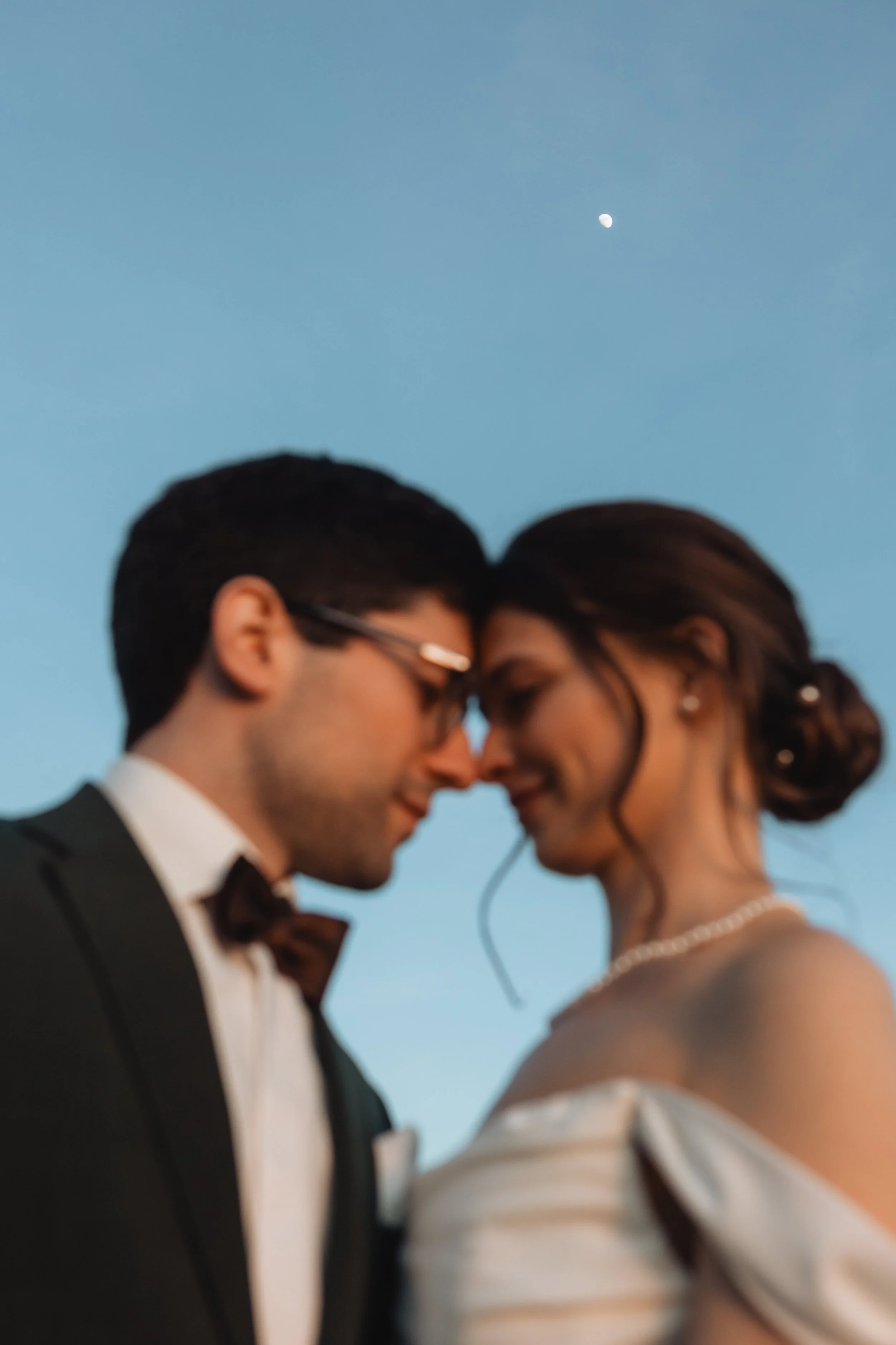 A close-up of a bride and groom touching foreheads with eyes closed during their wedding, with a blue sky and the moon visible in the background.