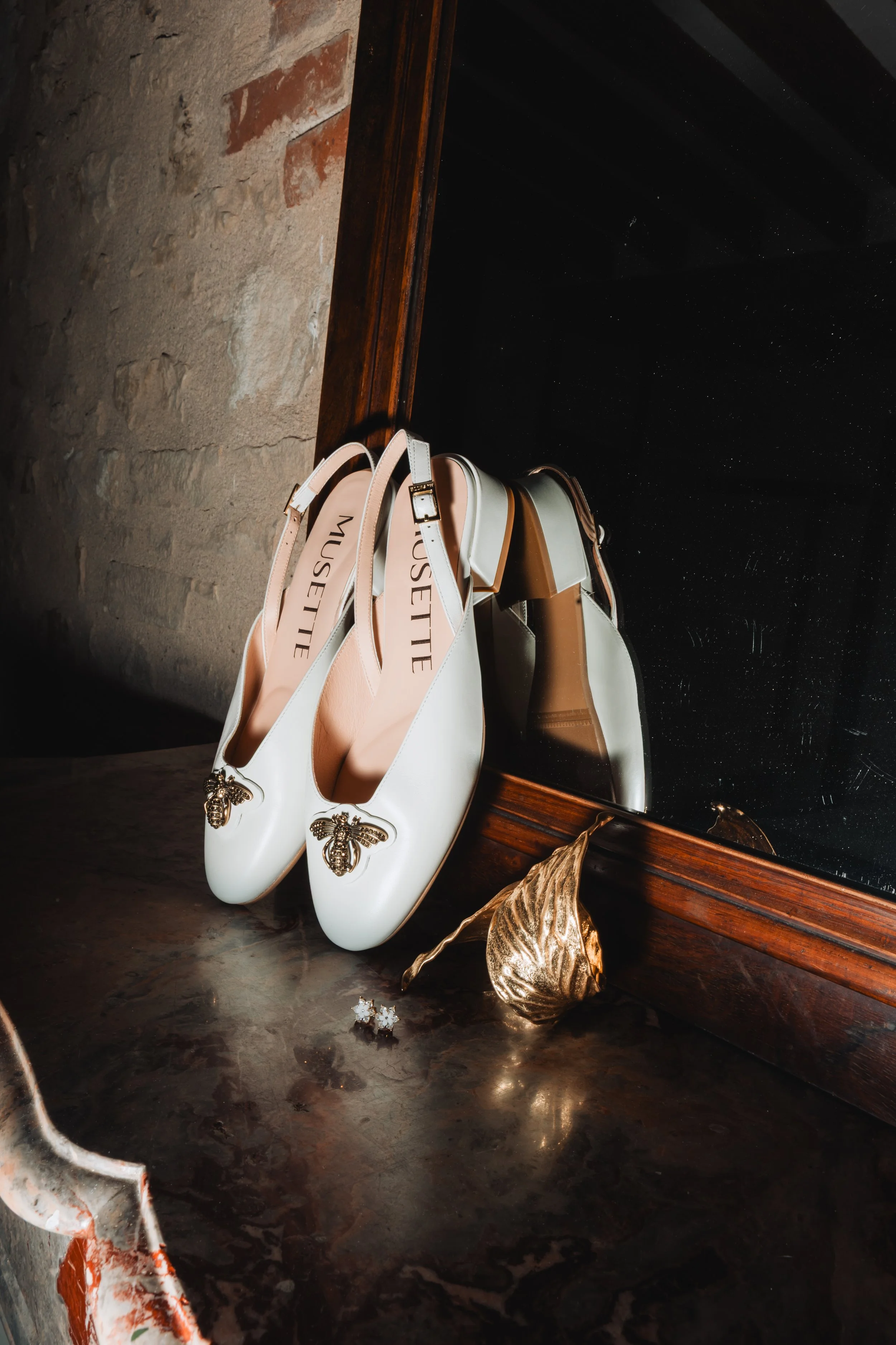 Pair of white shoes with bee-shaped embellishments on the toe, placed on a dark surface next to a reflective black mirror, with decorative golden leaves and a pair of flower-shaped earrings nearby.