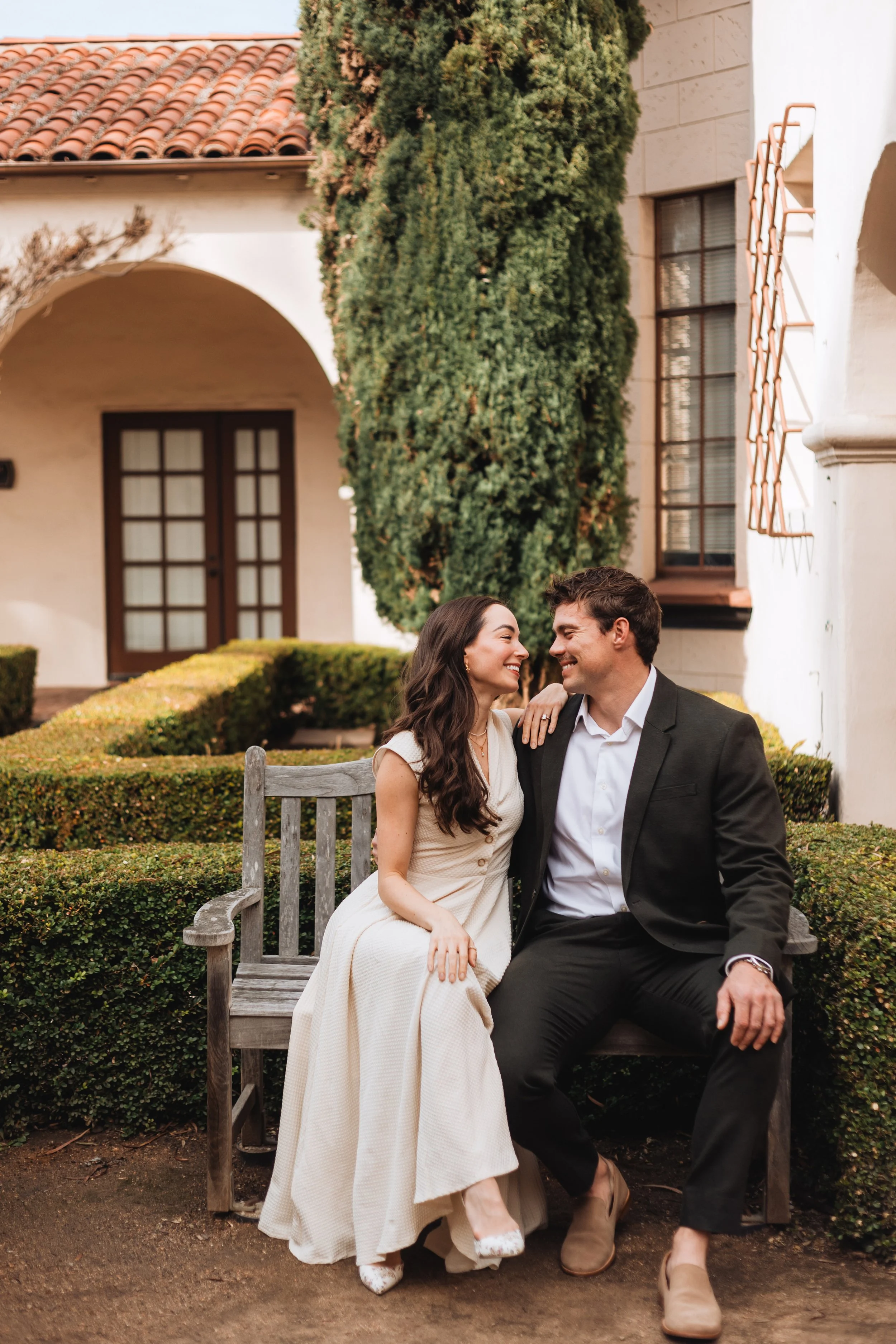 A couple sits on a wooden bench outside a building with Spanish-style architecture, including a red tile roof and arched entryway, surrounded by greenery. They are smiling at each other, dressed in formal attire.
