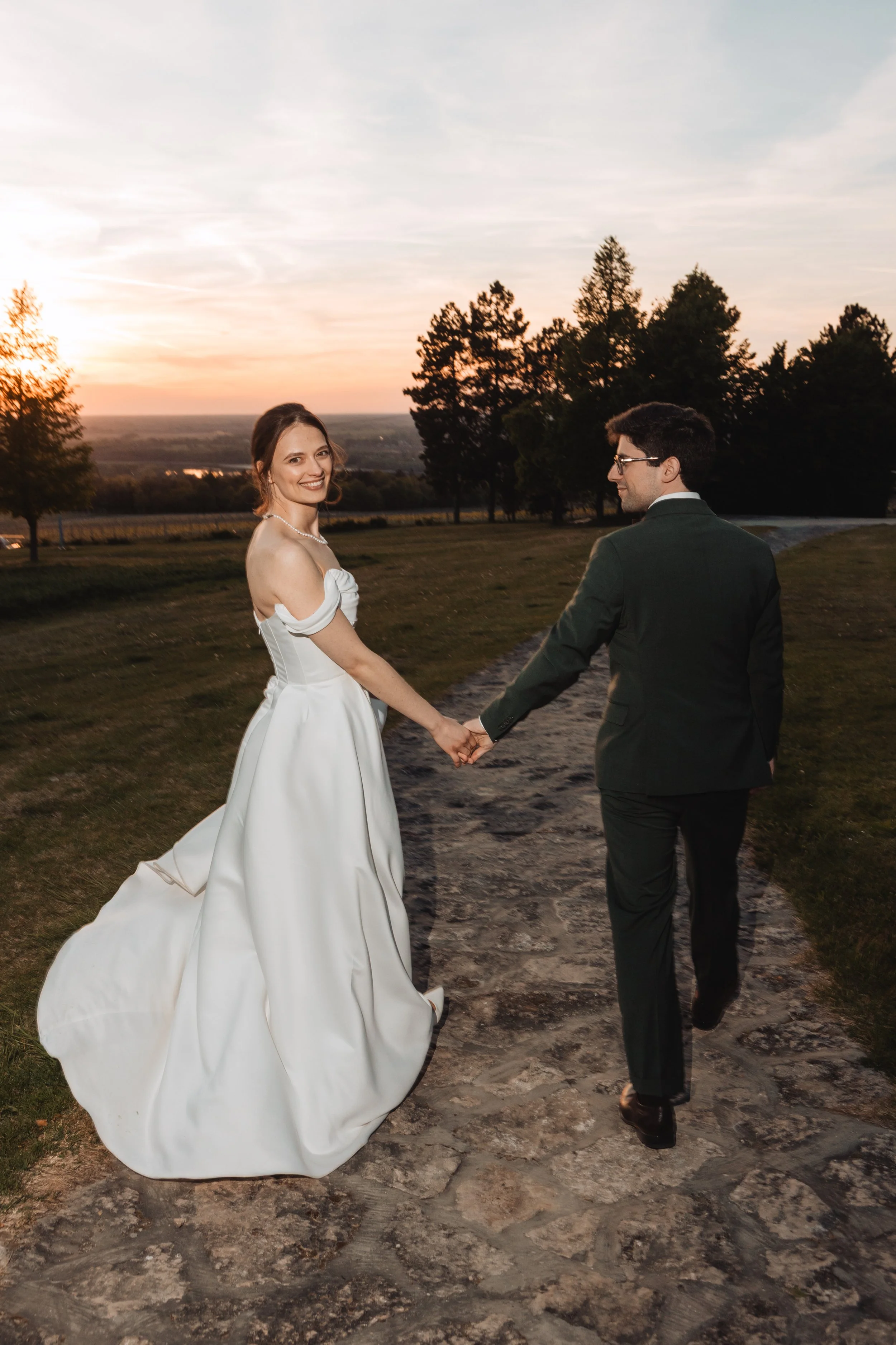 A bride and groom holding hands and walking on a stone path during sunset in a park, with trees and a view of the horizon in the background.