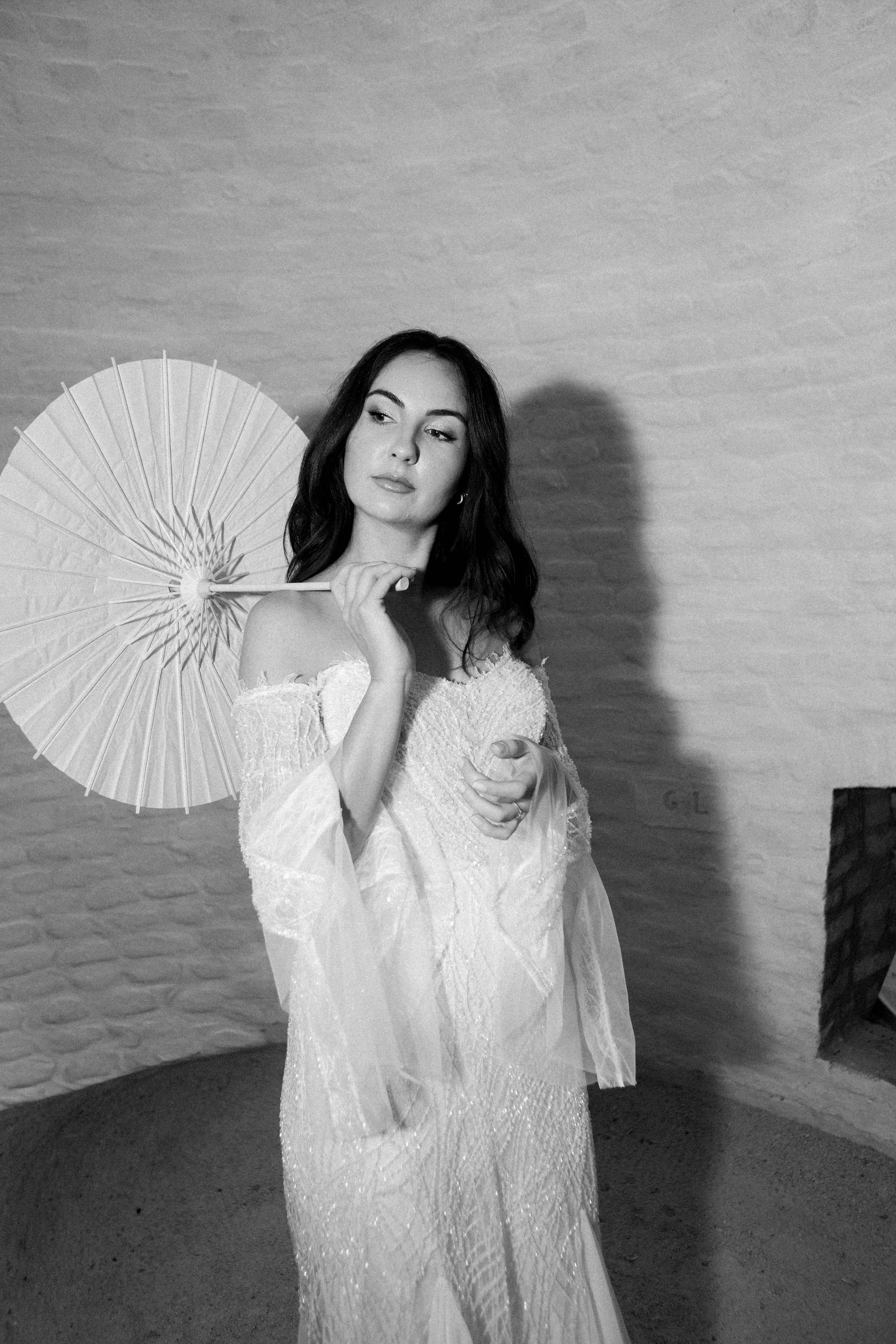 Black and white photo of a woman in a lace wedding dress holding a parasol behind her head, standing against a textured wall.