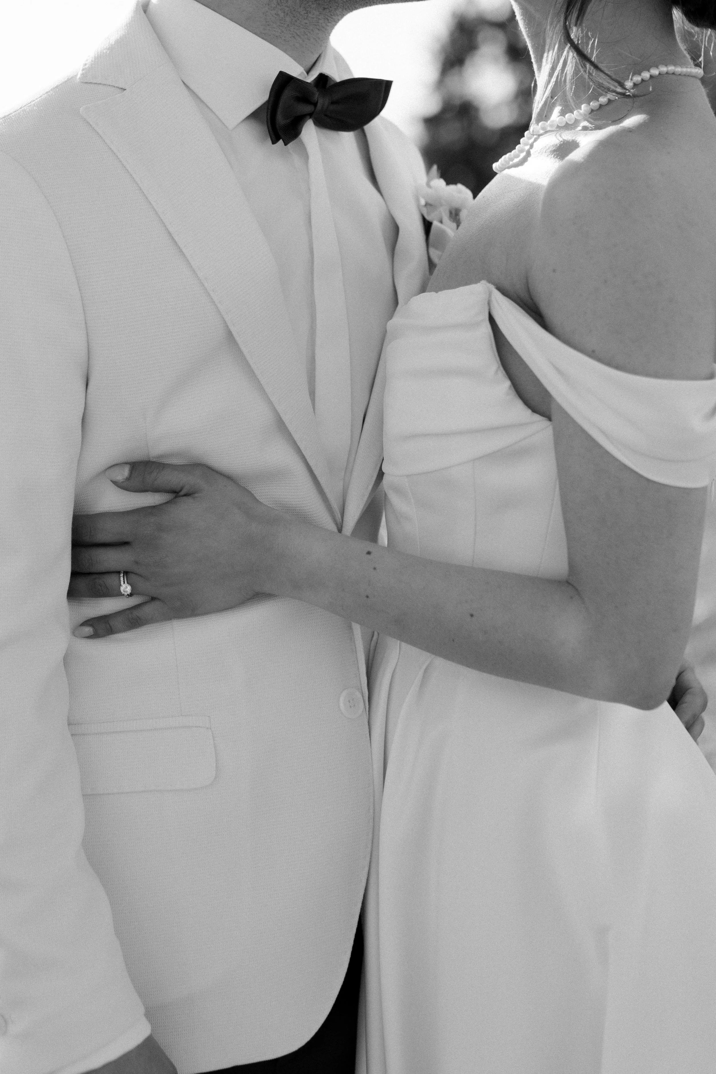 Close-up of a bride and groom, focusing on their torsos and arms, with the groom wearing a tuxedo with a bow tie and the bride in a strapless wedding gown, both standing closely together.