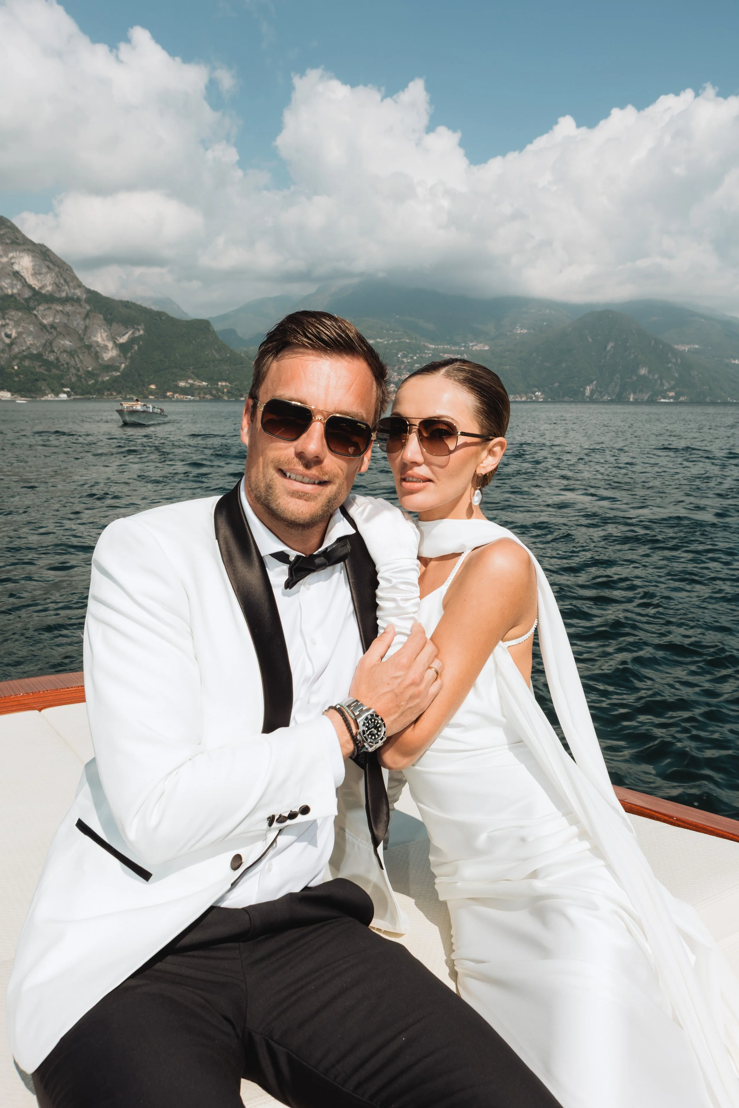 A couple dressed in formal tuxedo and white dress on a boat with mountains and water in the background