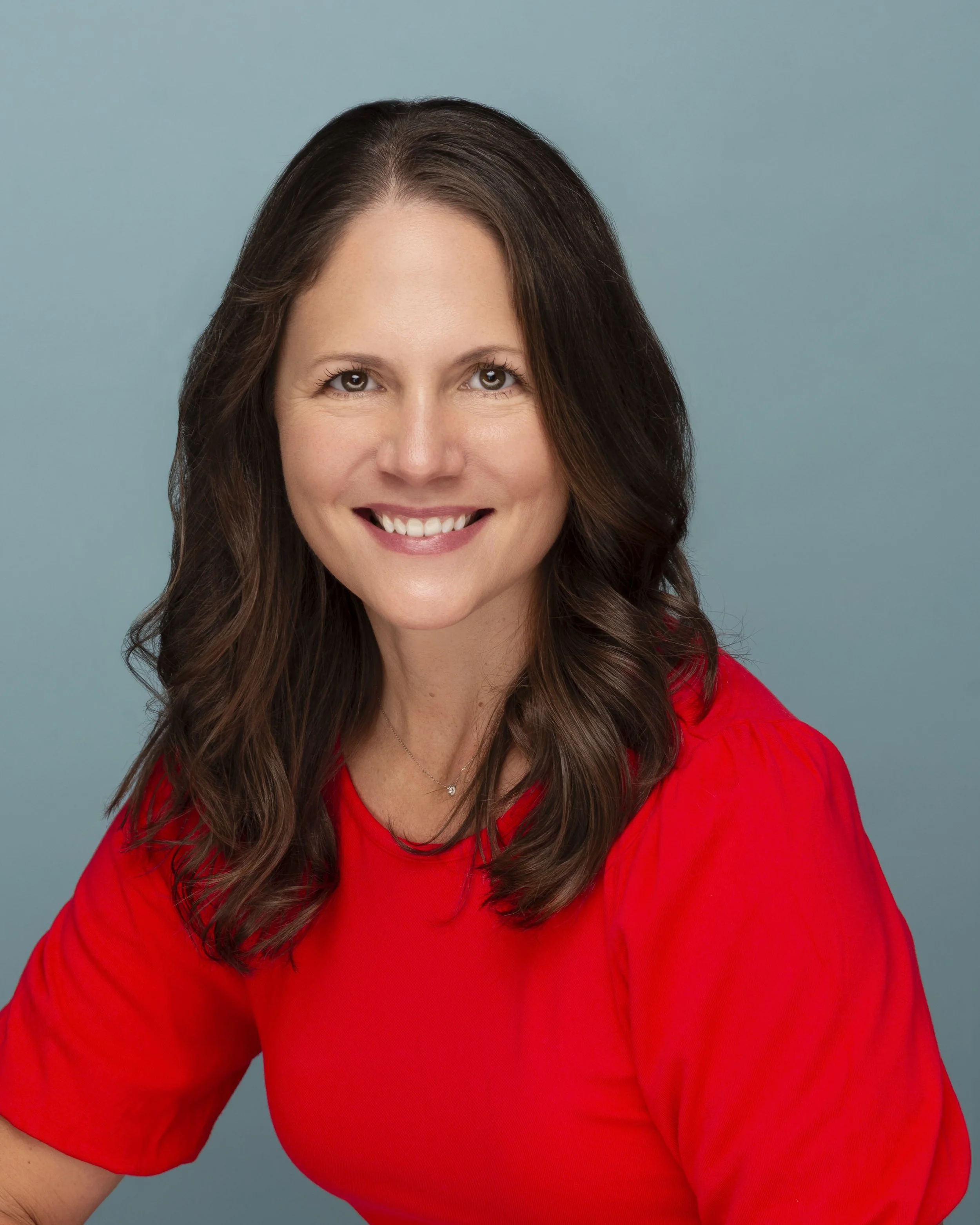 A woman with long brown hair, smiling, wearing a red top, against a light blue background.