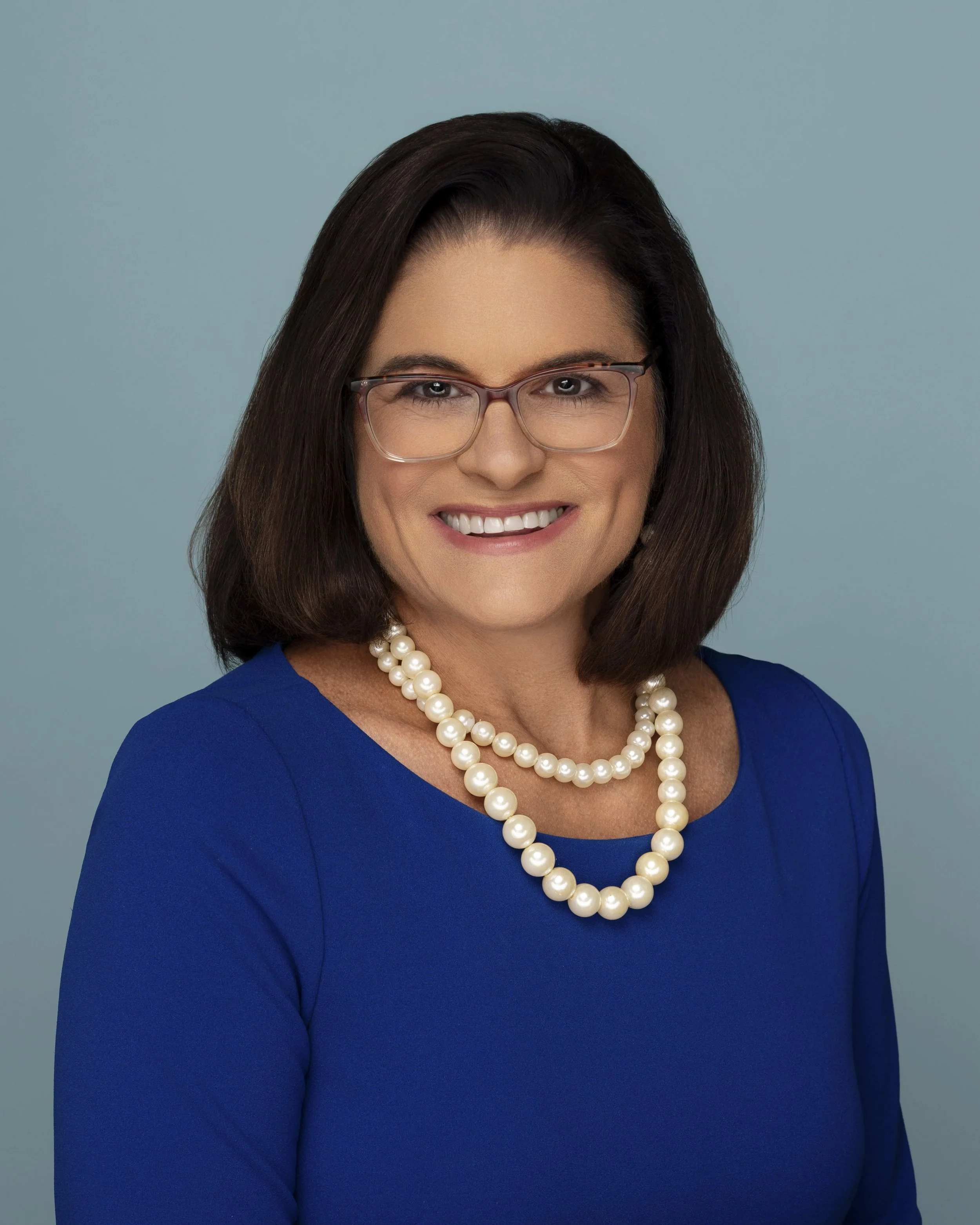 A woman with dark brown hair, glasses, and a friendly smile wearing a royal blue dress and two strands of pearls against a light blue background.