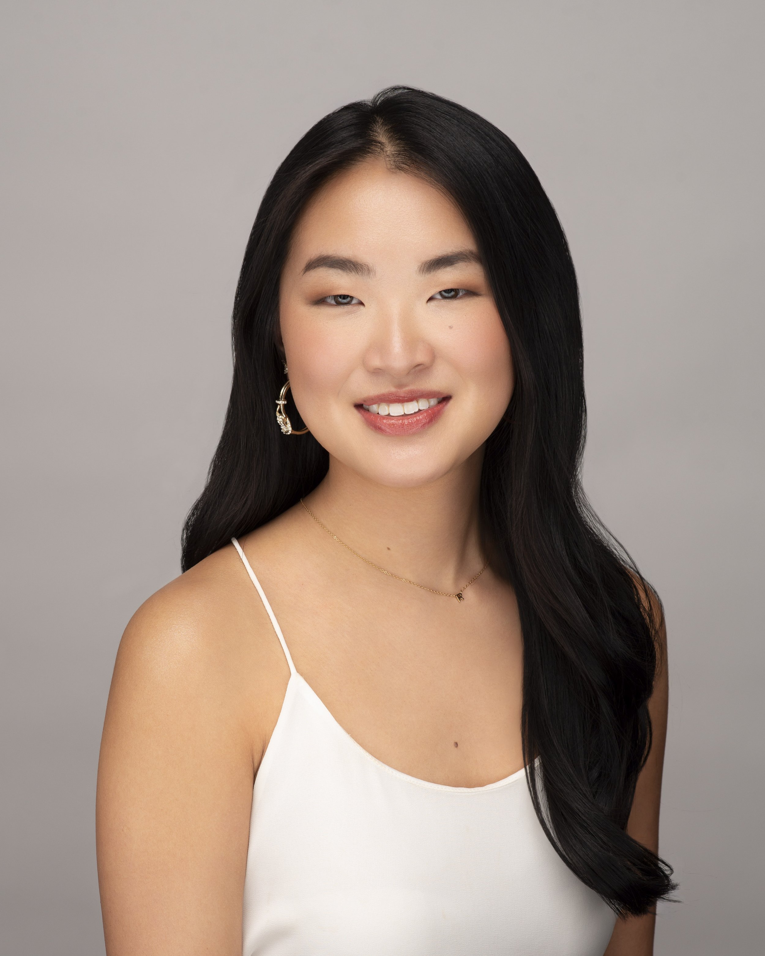 A young woman with long black hair, wearing a white spaghetti strap top, gold hoop earrings, and a delicate gold necklace, smiling at the camera against a neutral gray background.