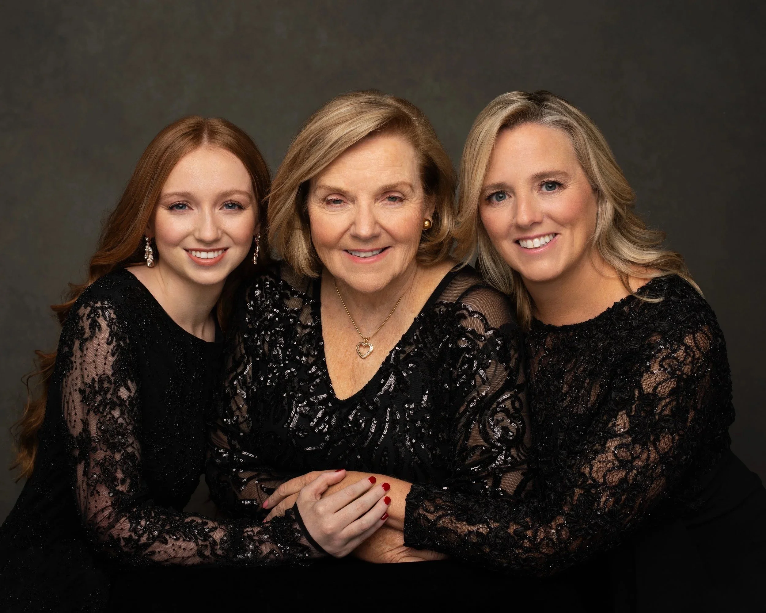 Three women, a young woman, an older woman, and a middle-aged woman, dressed in black lace clothing, smiling and posing together against a dark background.