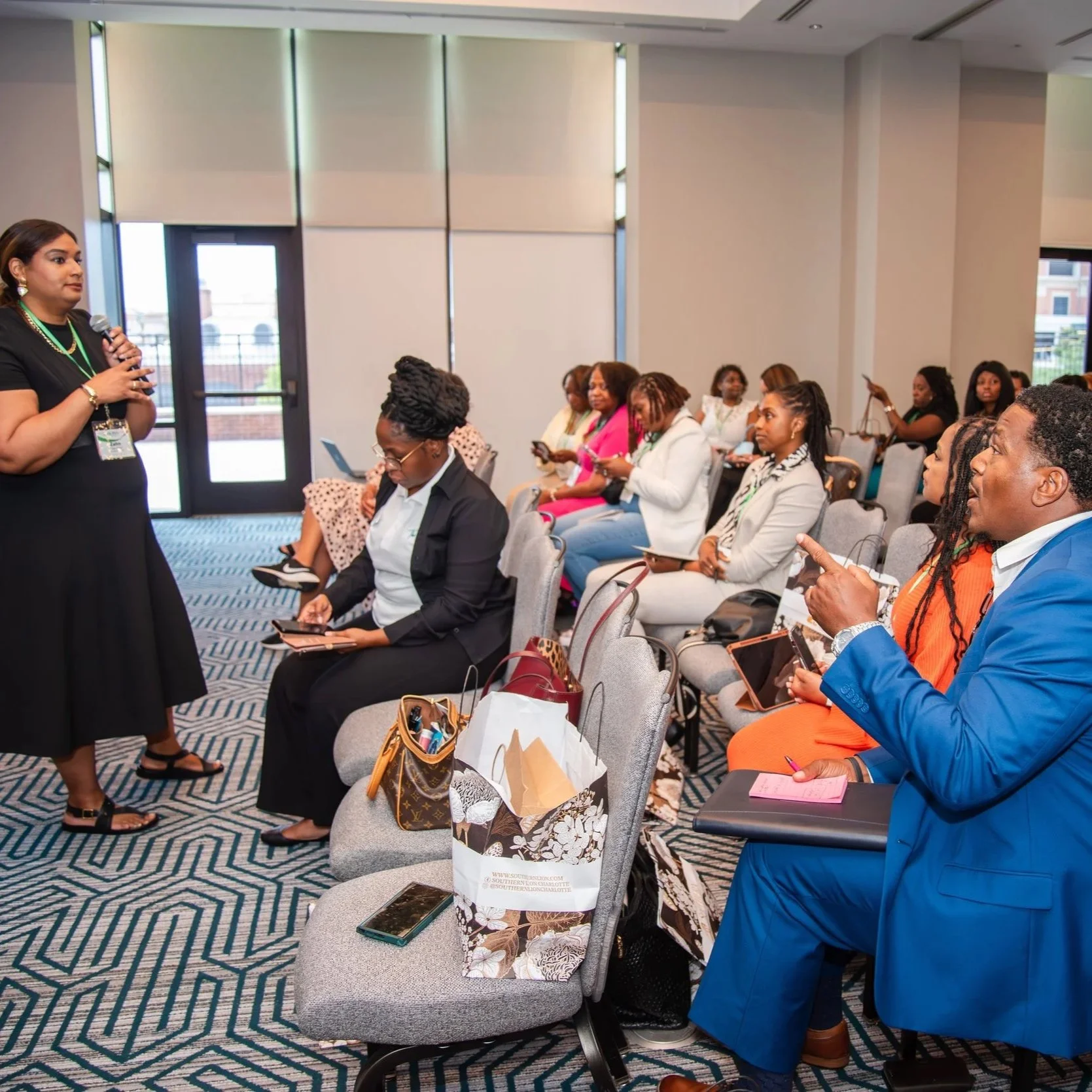 A woman in a black dress giving a presentation at a conference, with an audience of men and women seated and listening attentively in a modern conference room.