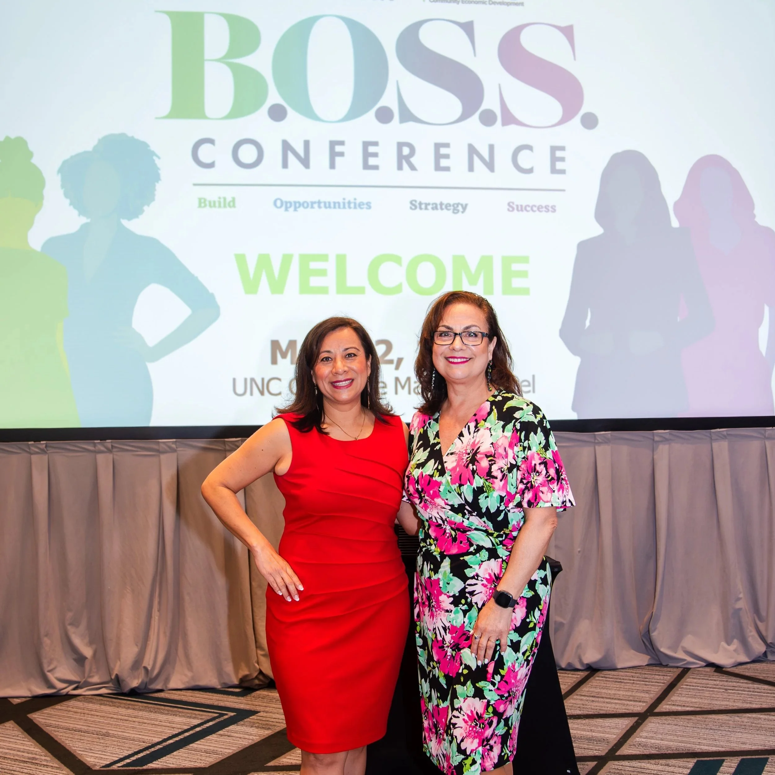 Two women standing together at the B.O.S.S. Conference, smiling for the camera. Background shows a welcome message on a screen with silhouettes of people and colorful text.