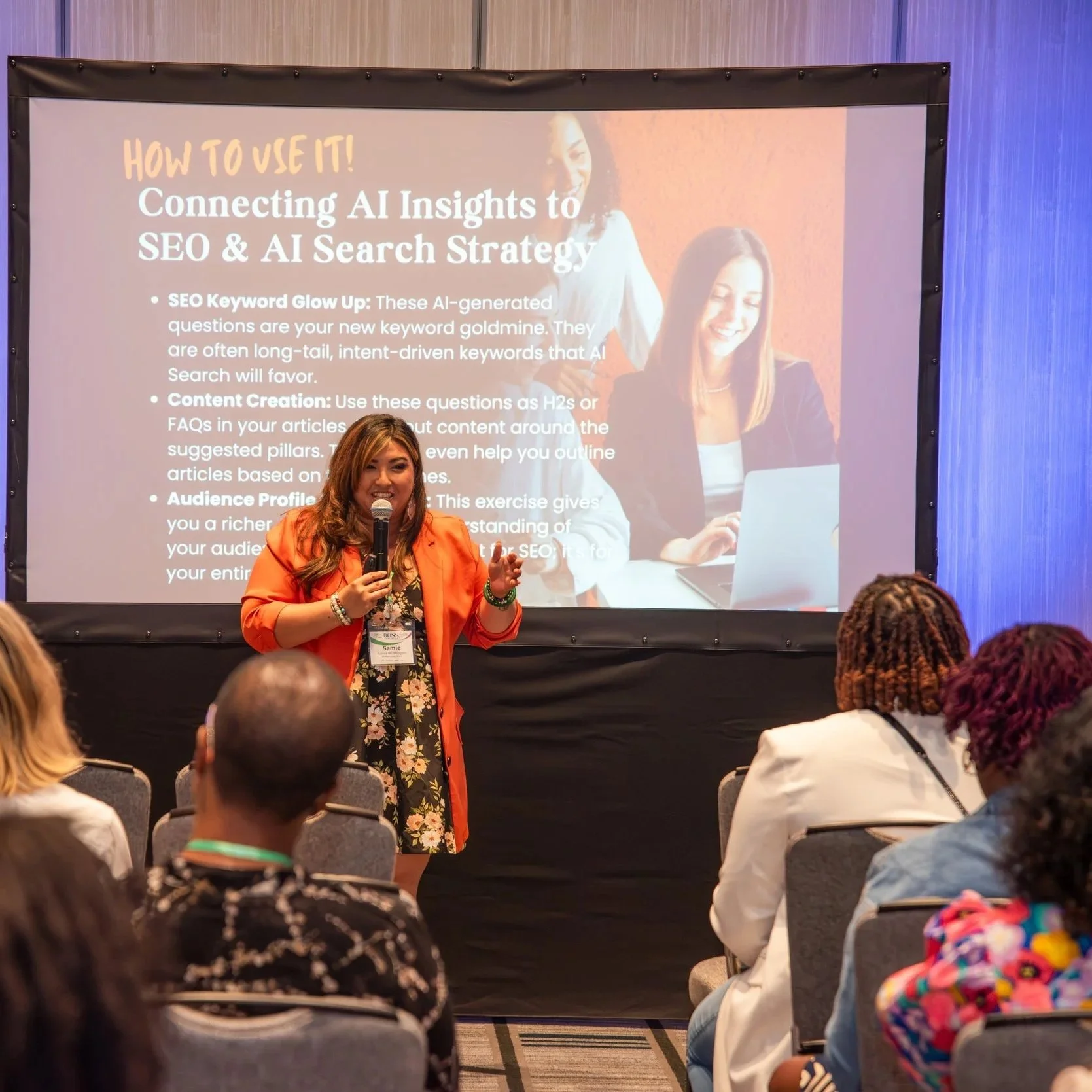 A woman in an orange blazer presents at a conference, holding a microphone, in front of a large screen displaying a slide about AI insights and SEO strategies. Audience members sit and listen.