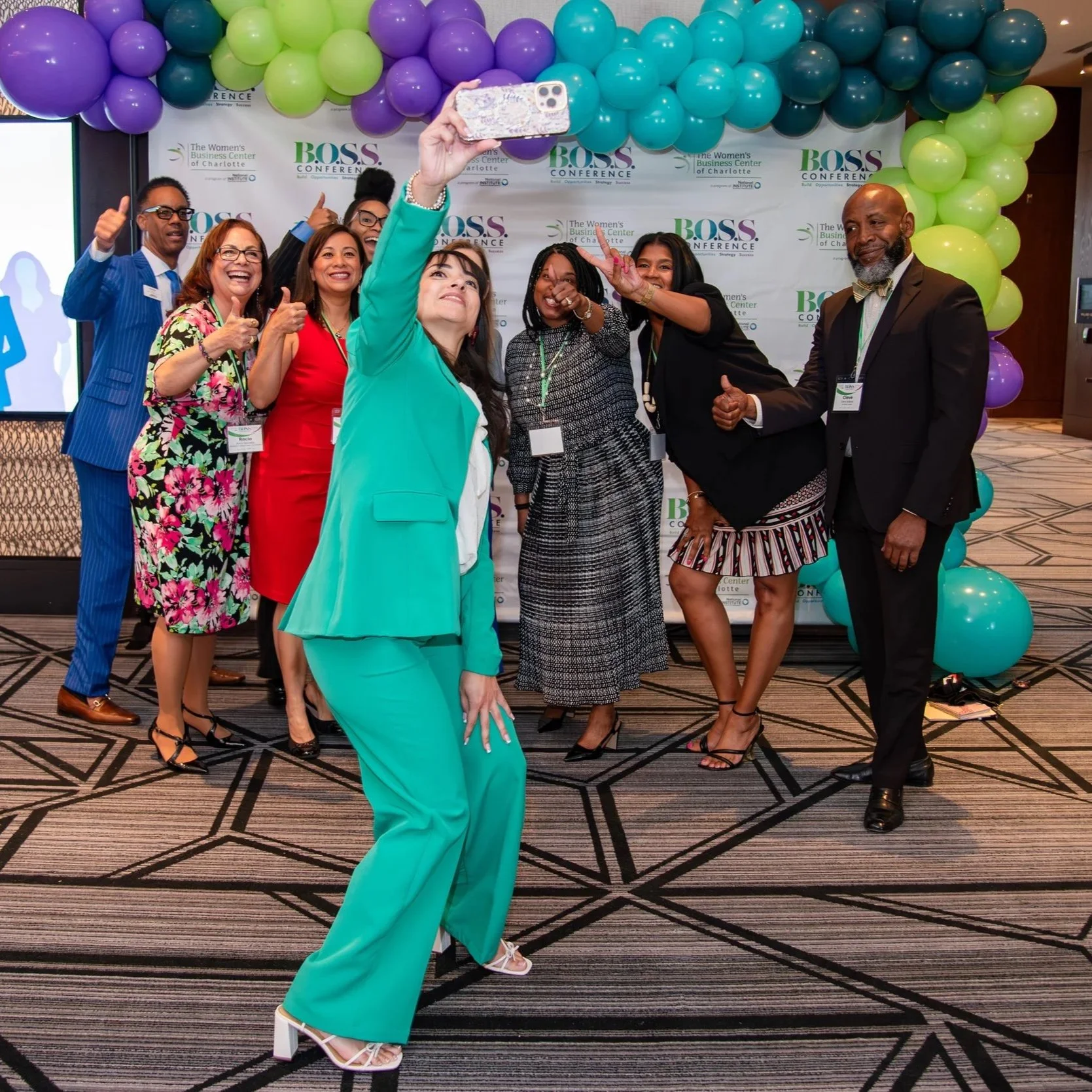 Group of diverse women and a man at a conference taking a selfie in front of a decorated backdrop with balloons and the conference logo.