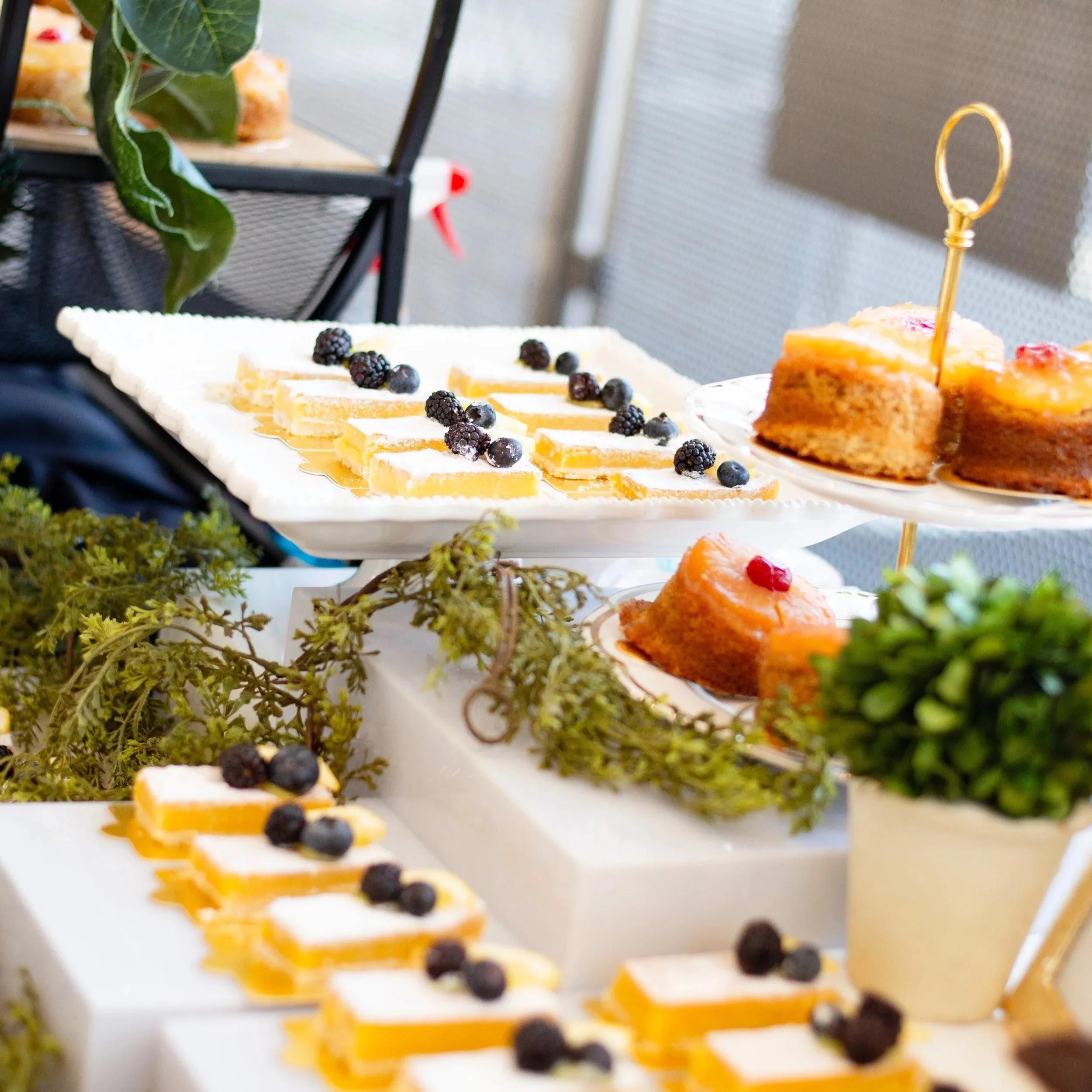 Assorted small cakes and pastries on white tiered serving platters, decorated with berries, with greenery and plants around, at an outdoor event or gathering.