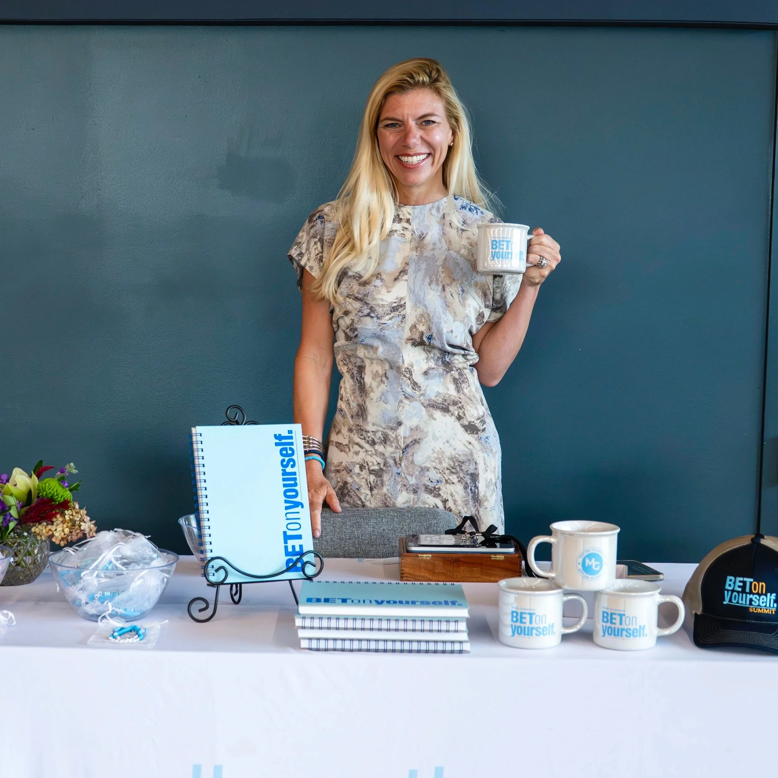 A woman with blonde hair smiling and holding a mug, standing behind a table with promotional items, mugs, notebooks, and flowers against a blue wall.