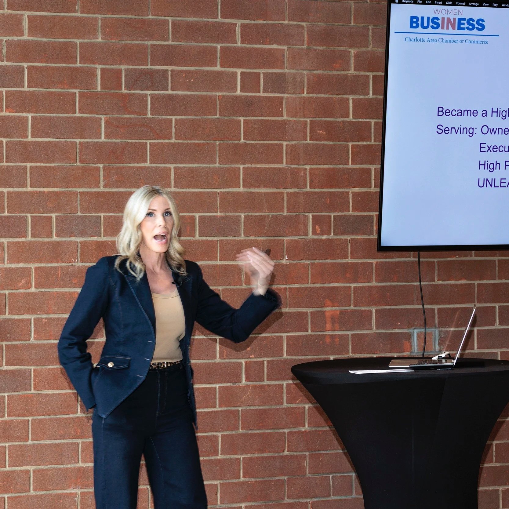 A woman with blonde hair, wearing a black blazer and beige top, giving a presentation in front of a brick wall. A large screen to her right displays a slide with the heading 'Women Business' and the Charlotte Area Chamber of Commerce logo.