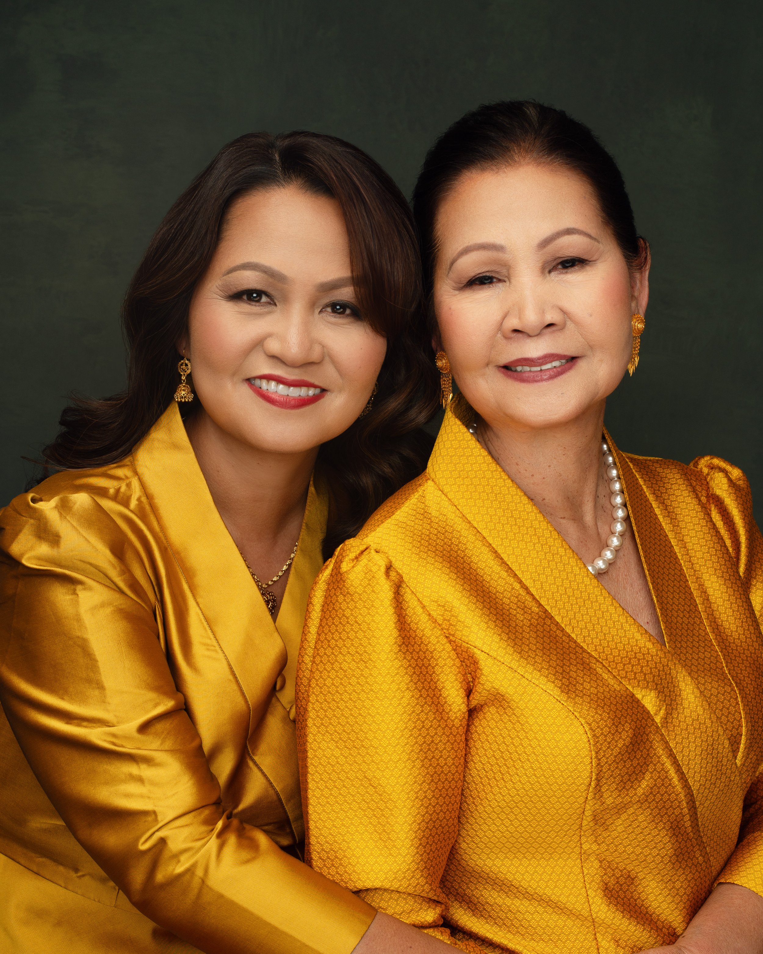 Two women dressed in yellow silk outfits, wearing pearl and gold jewelry, smiling for the camera against a green background.