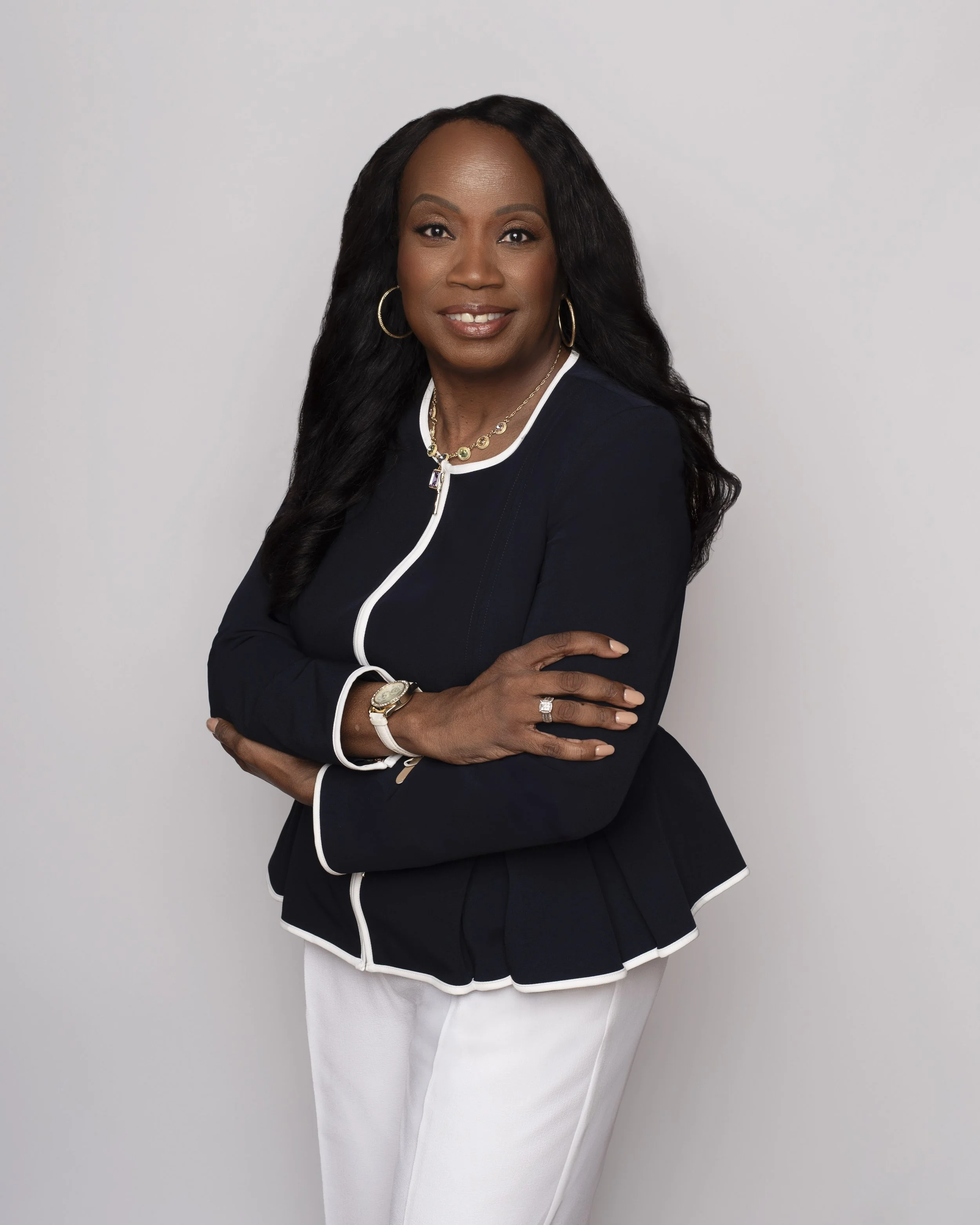 A woman with long dark hair, wearing a black jacket with white piping and white pants, standing against a white background with her arms crossed, smiling at the camera.