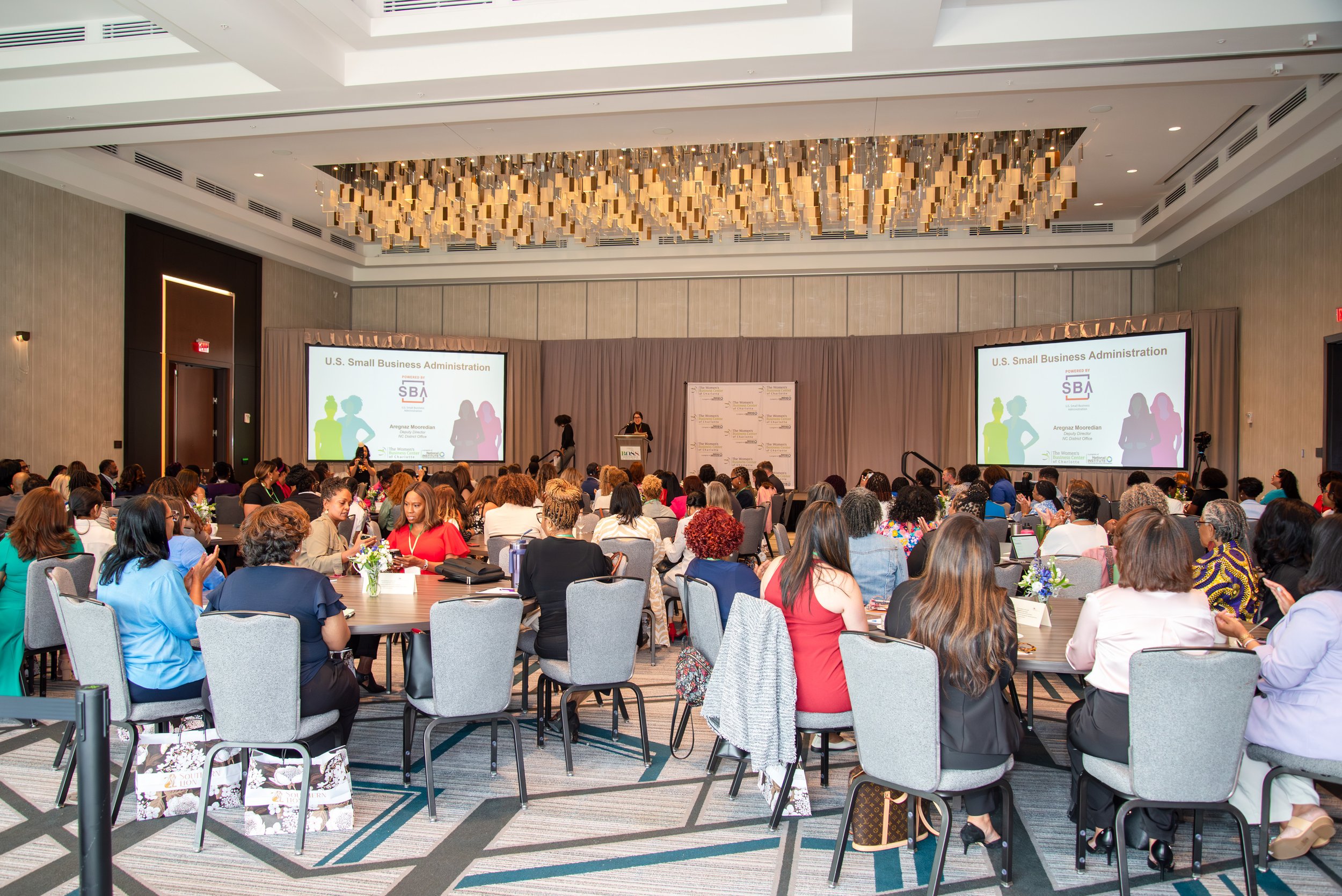 A large conference room filled with people attending a presentation on U.S. Small Business Administration. There are two large screens displaying the presentation and a speaker at a podium at the front of the room.