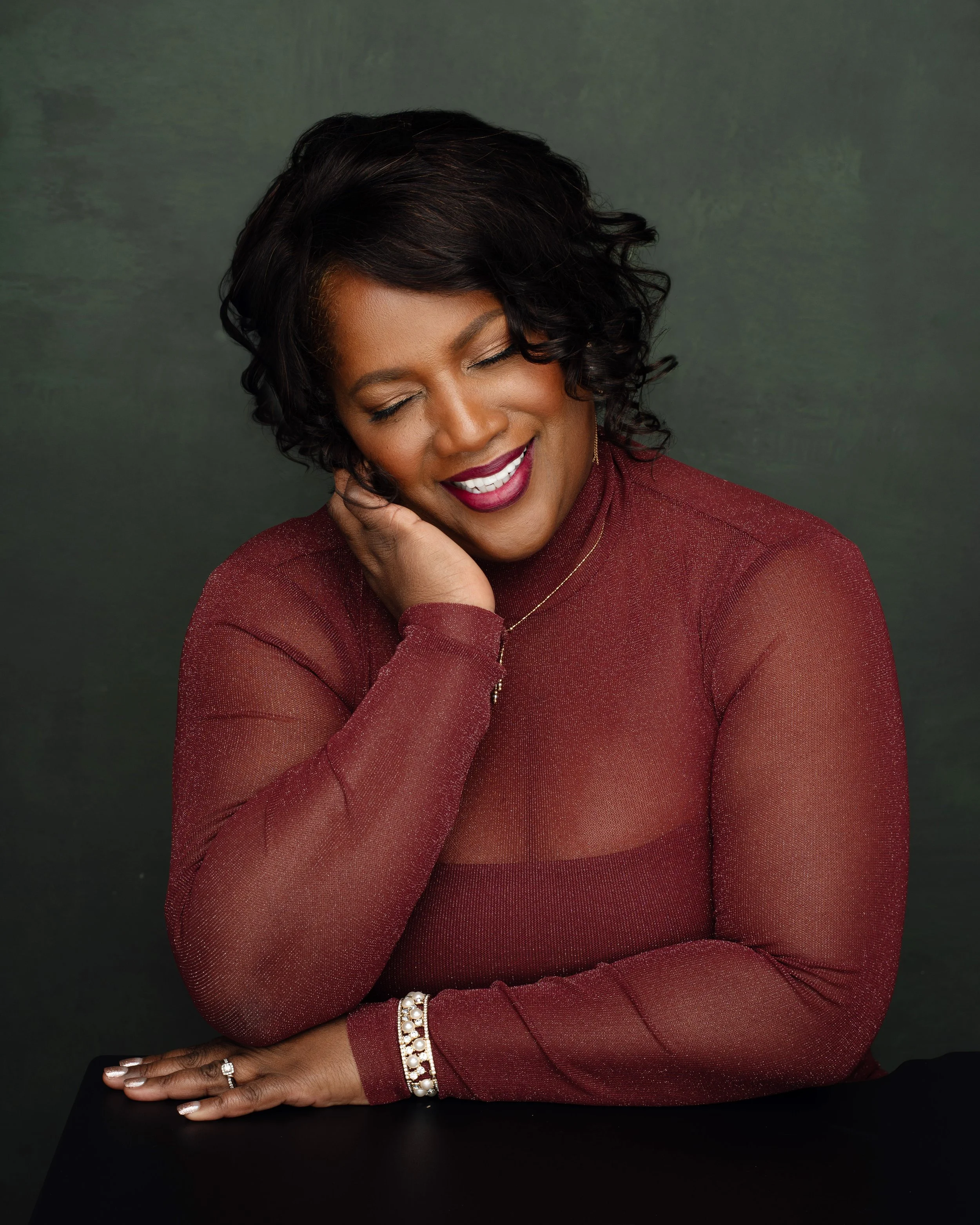 A woman with dark, curly hair, wearing a sheer, long-sleeved, red top, smiling with eyes closed, with one hand resting on her face and the other on a black surface, against a dark green background.