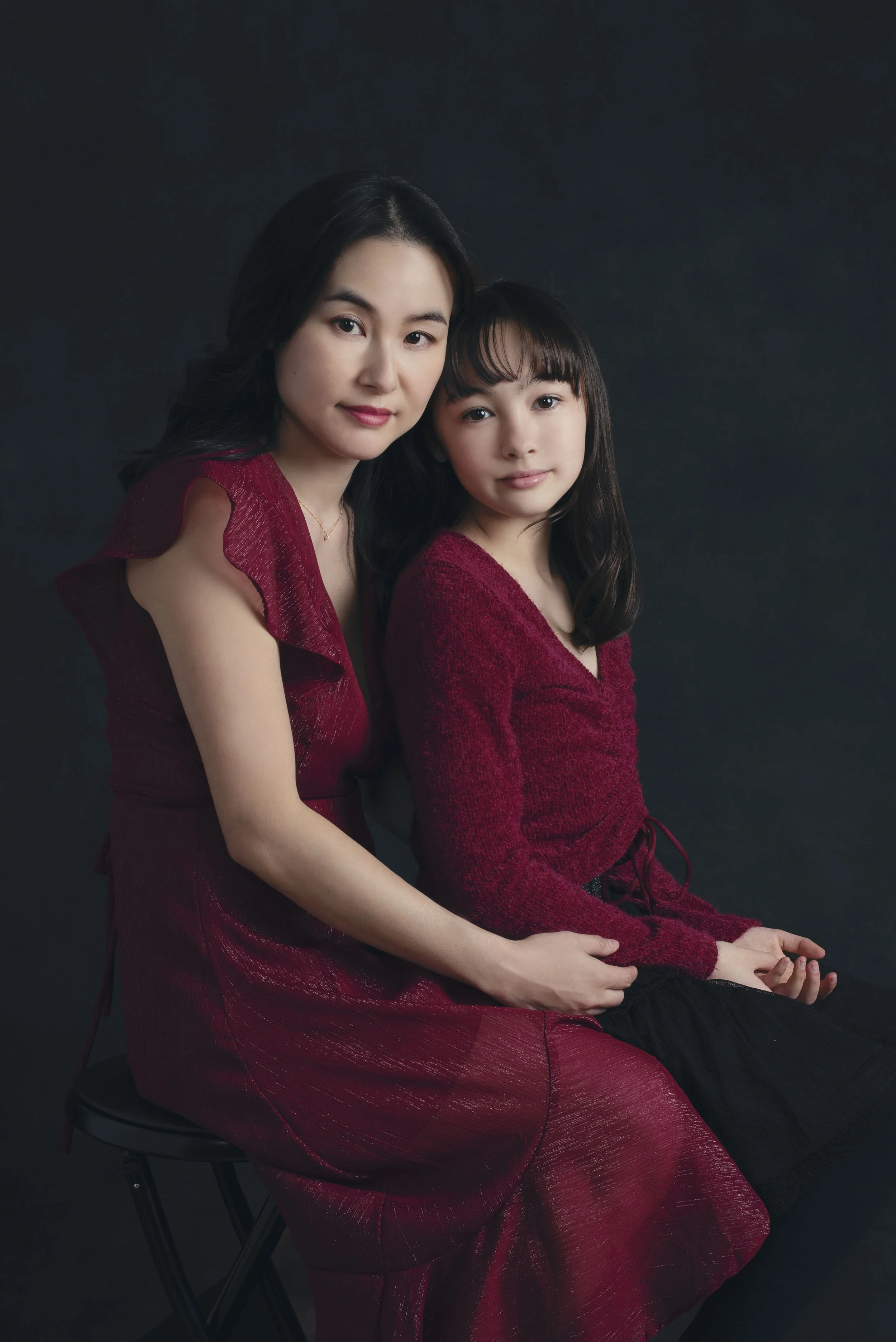 A woman and a young girl sitting together against a black background. Both are wearing red outfits and looking at the camera.