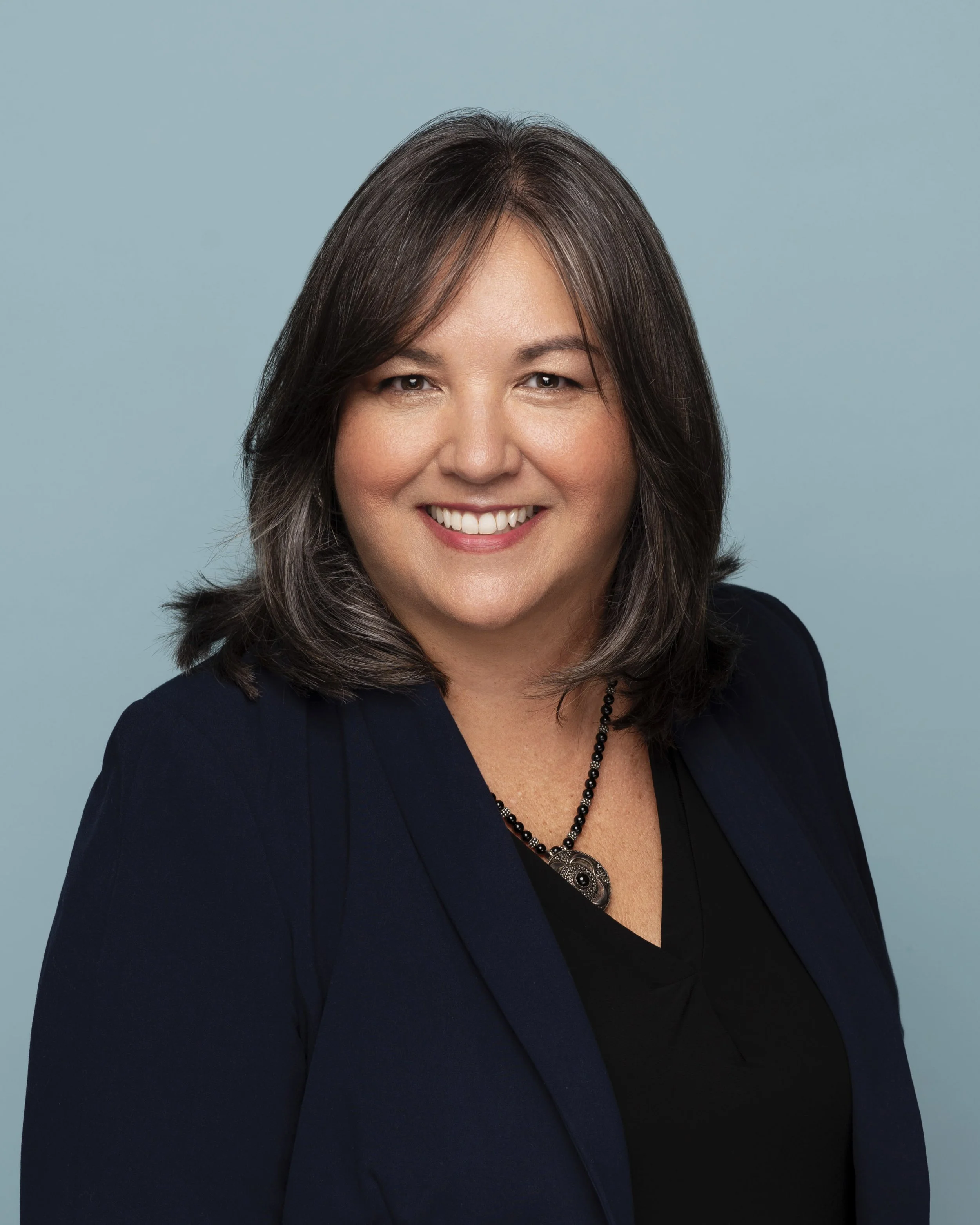 Headshot of a woman with shoulder-length dark brown hair, wearing a dark blazer, a black top, and a necklace, against a light blue background.