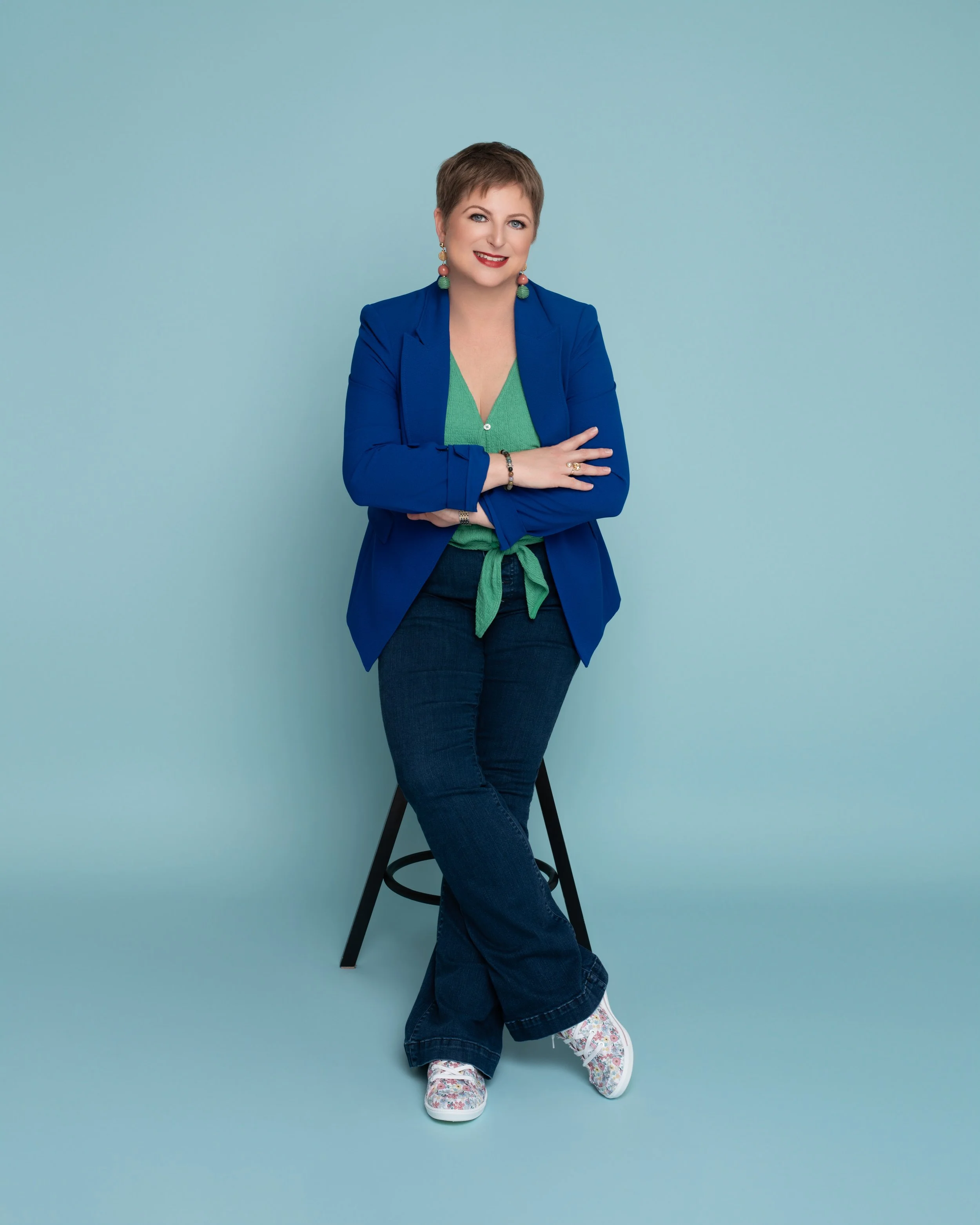 A woman with short brown hair, wearing a blue blazer over a green shirt, dark jeans, and white floral sneakers, sitting on a black stool against a light blue background and smiling.