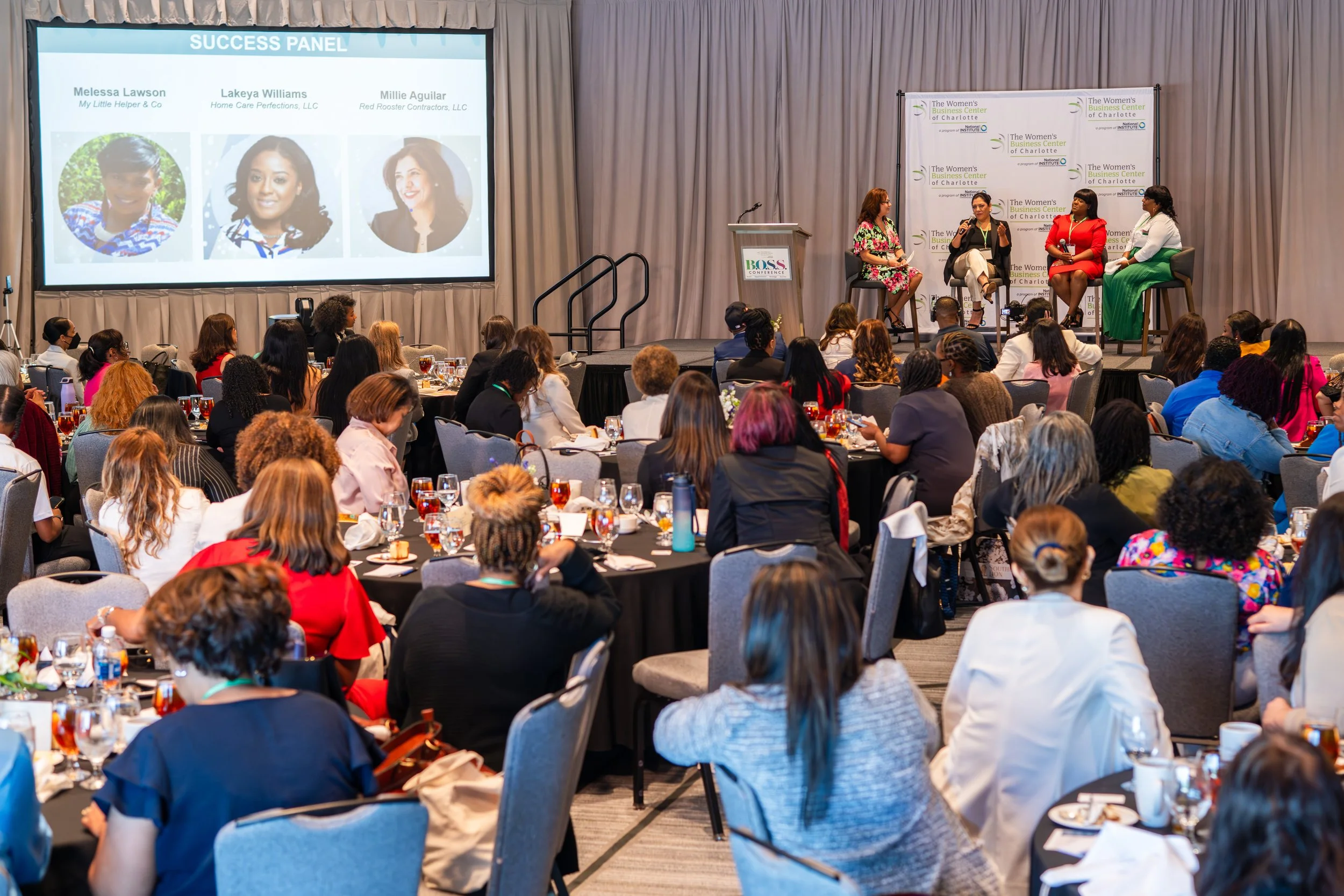 A large conference room filled with women seated at round tables watching a panel discussion on stage. The stage has four women, one speaking into a microphone, with a backdrop and a podium. A large screen displays panelist names and photos.