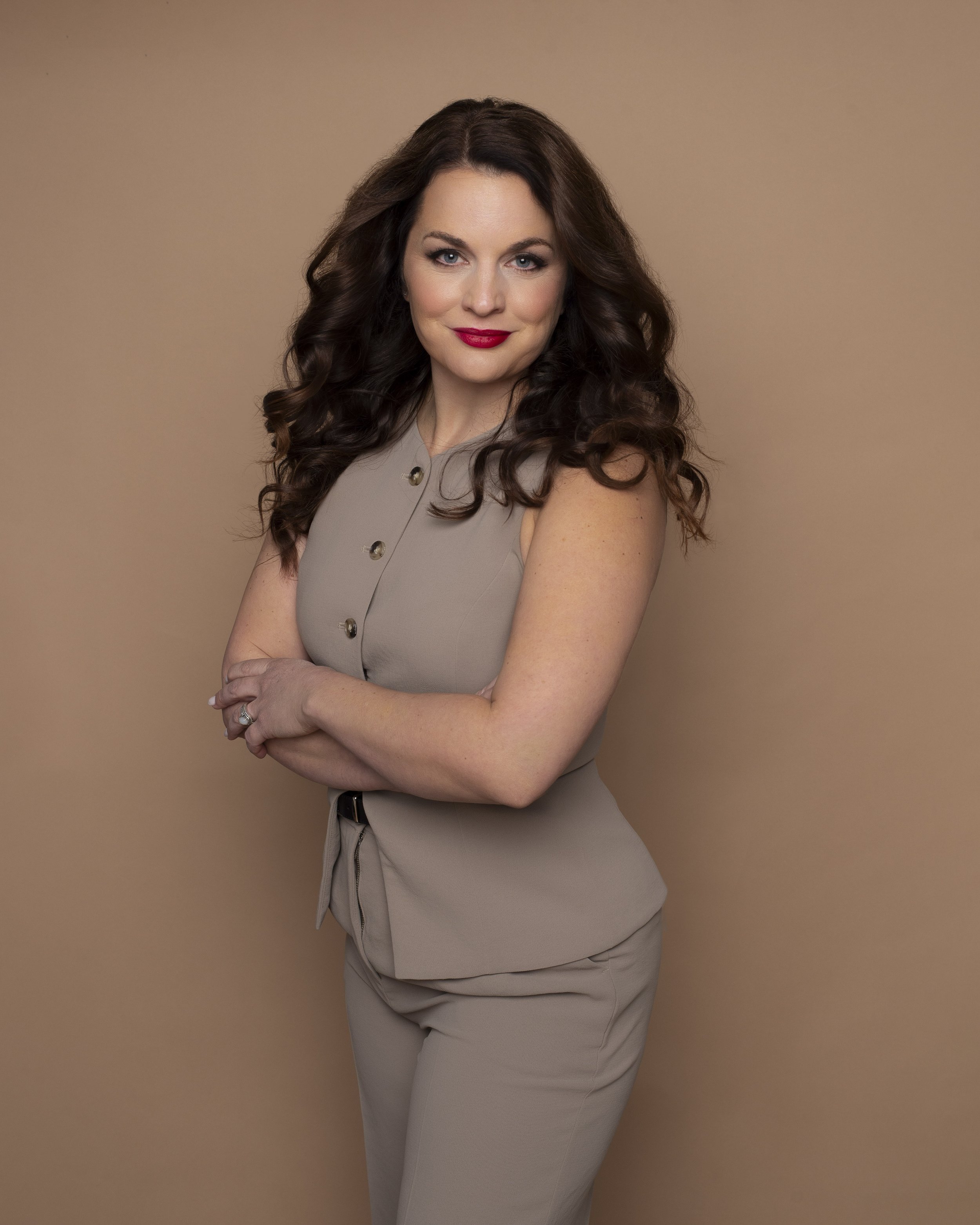 A woman with long, dark, wavy hair, wearing a beige sleeveless top and matching pants, standing against a plain beige background.