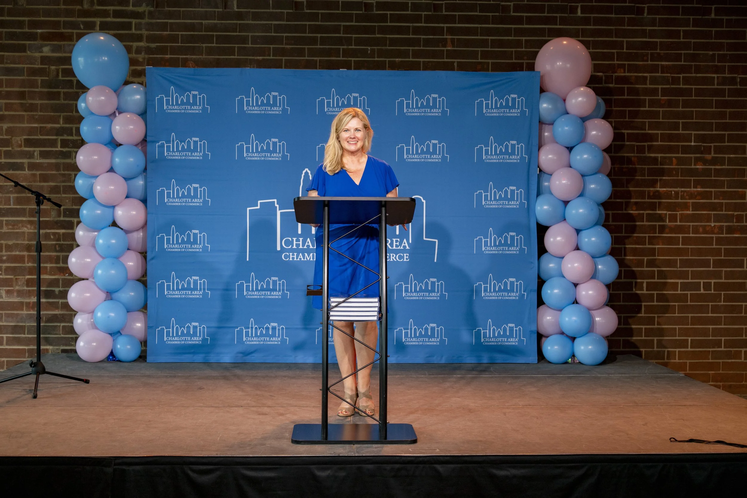 A woman stands at a podium on a stage decorated with a blue backdrop and pink and blue balloon columns. The backdrop has the Charlotte Area Chamber of Commerce logo repeated across it.